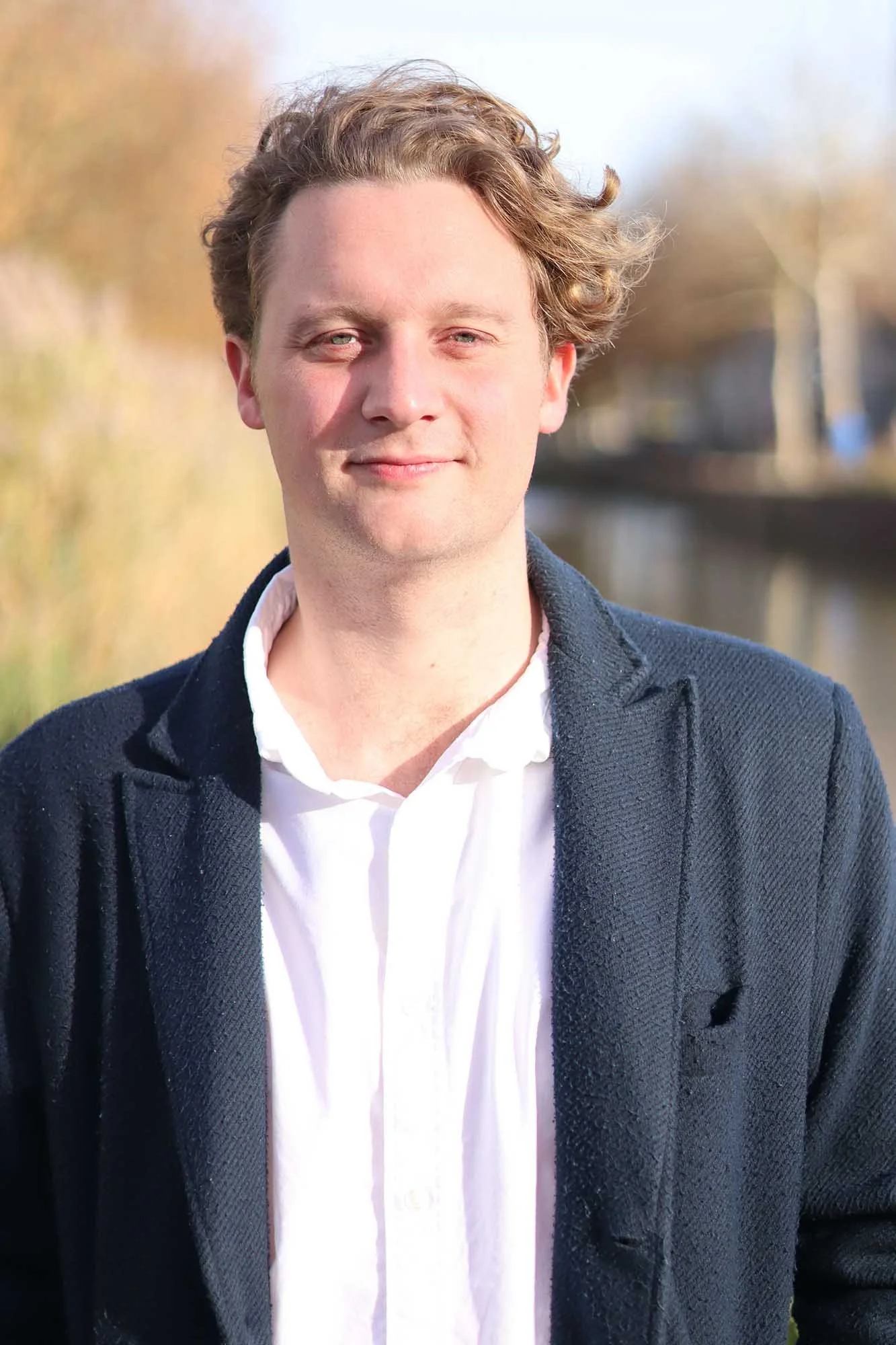 Portret van een jonge man met lichtbruin, golvend haar, gekleed in een witte blouse en een donkerblauw blazer, staand buiten met een waterkanaal en bomen op de achtergrond.