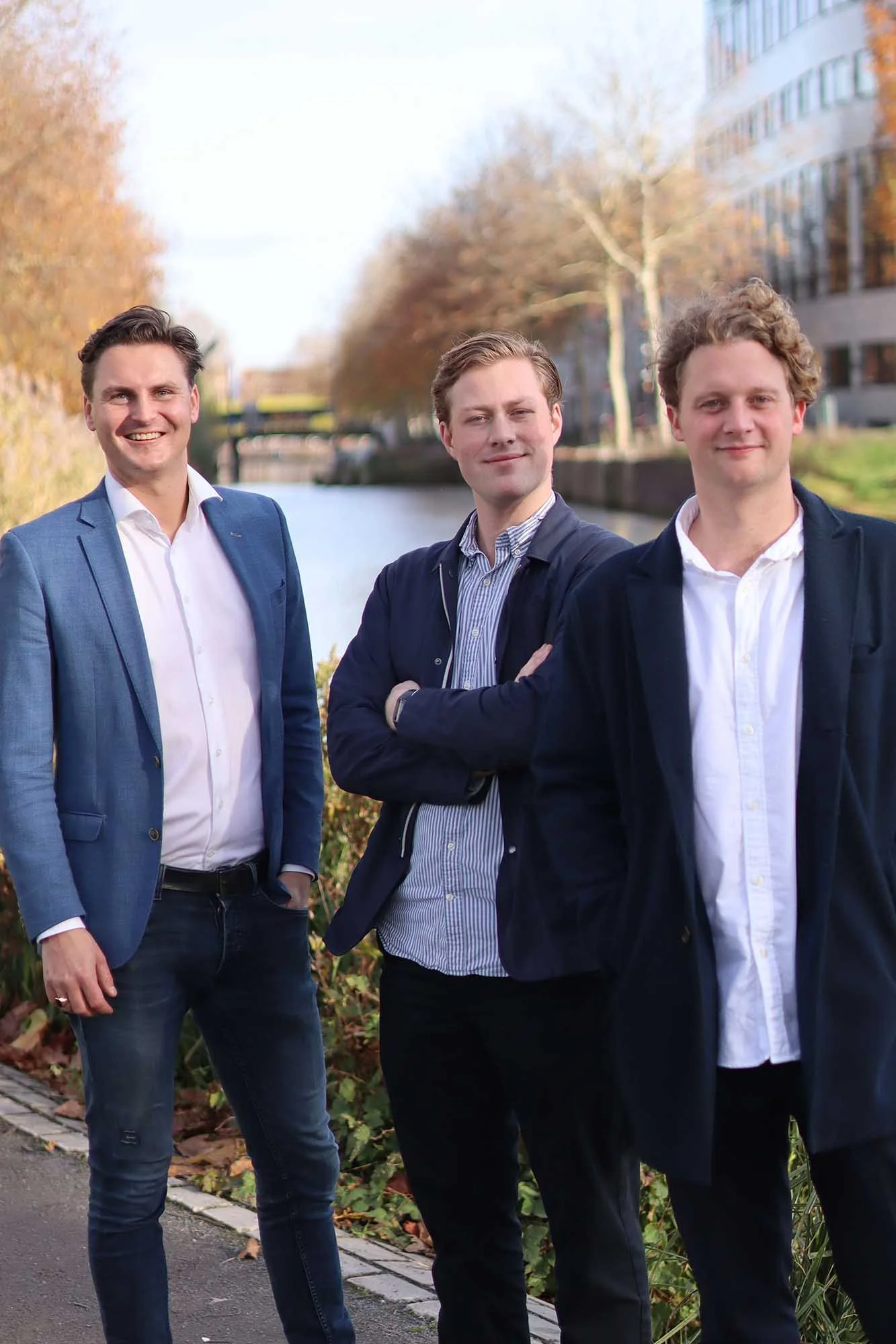 Drie jonge mannen in zakelijke kleding staan langs een kanaal met gebouwen en bomen op de achtergrond.
