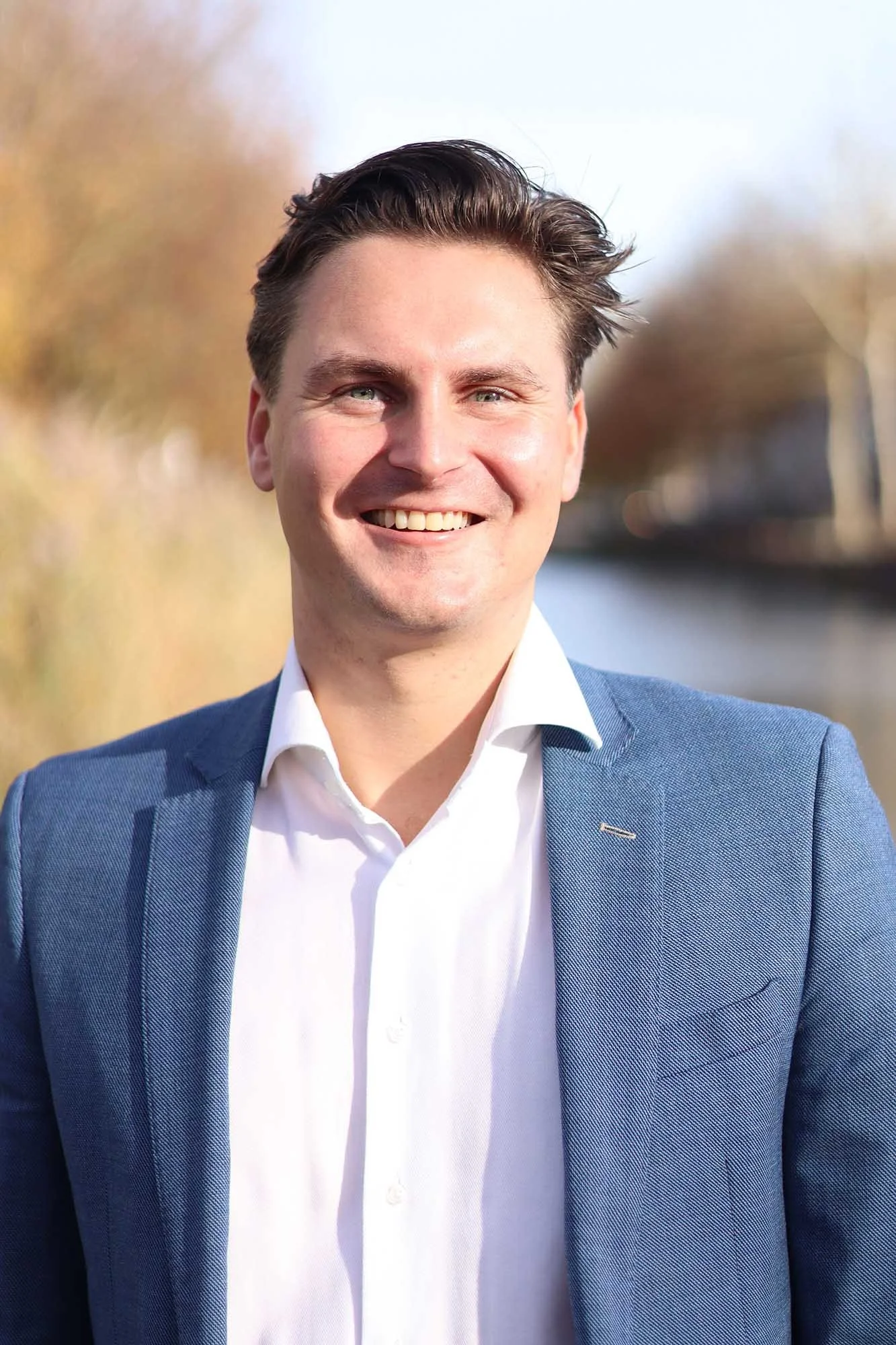 Lachende man in zakelijke kleding buiten, met water en bomen op de achtergrond.