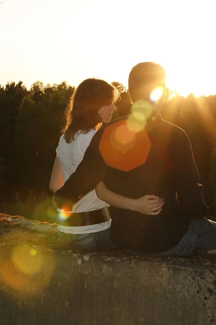 Ein Mann und eine Frau sitzen eng beieinander auf einer Mauer bei Sonnenuntergang, umgeben von Bäumen, mit Lichtreflexen und Sonnenstrahlen, die eine romantische Stimmung erzeugen.