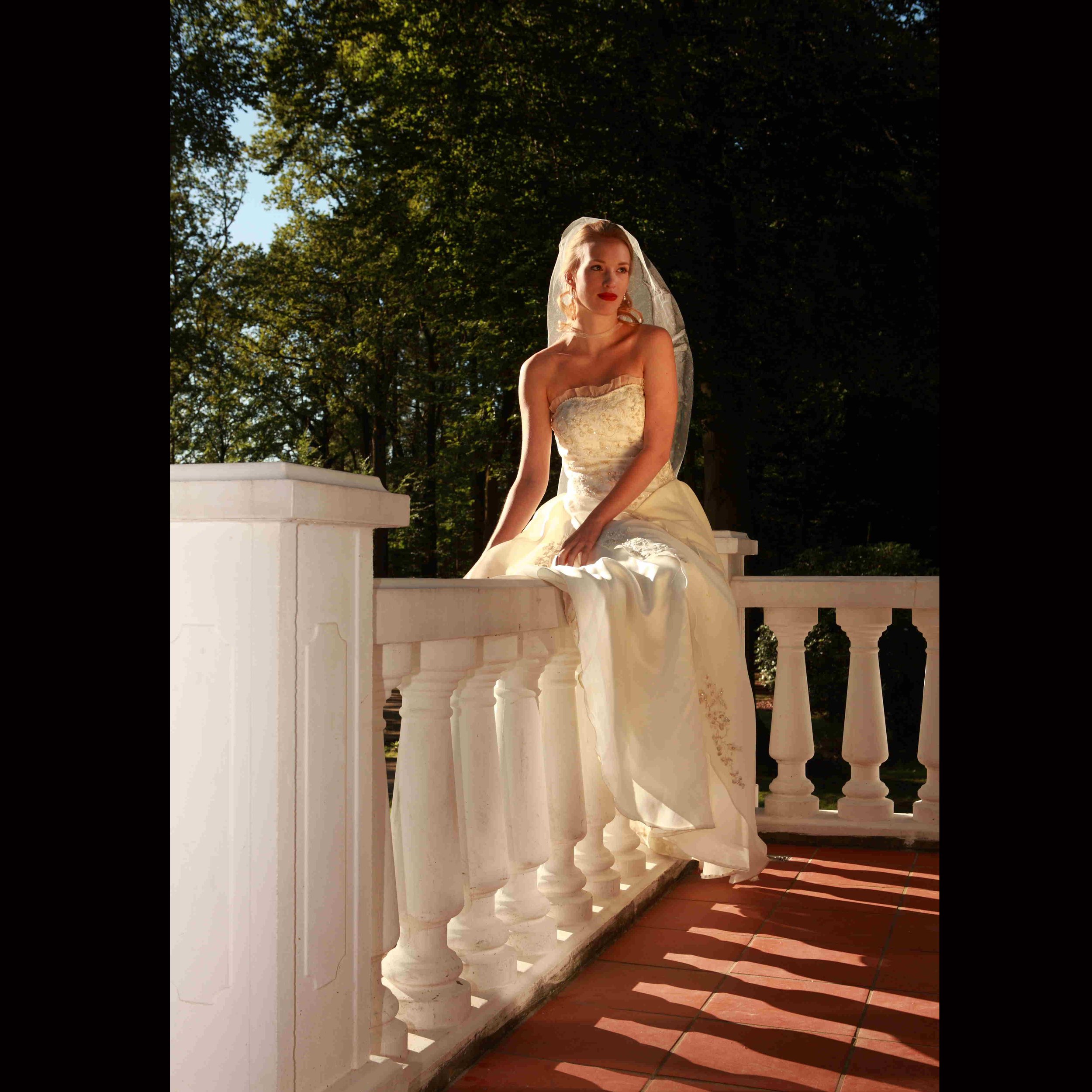 Eine Frau in einem weißen Brautkleid mit Schleier sitzt auf einem weißen Geländer im Freien, umgeben von grünen Bäumen, Sonnenlicht und Schatten auf terrakottafarbenen Fliesen.