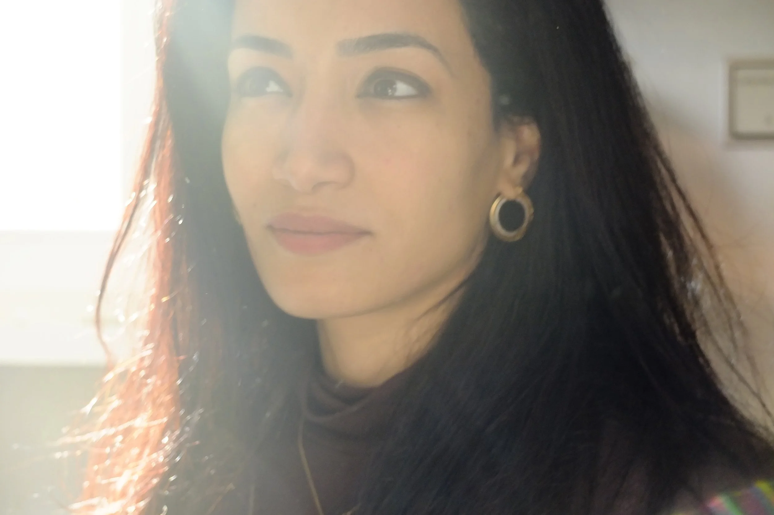 Nahaufnahme eines Frauen Gesichts, lachend, mit goldenen Ohrringen, dunklem Haar und make-up, im Hintergrund unscharf, mit Lichtreflexen.
