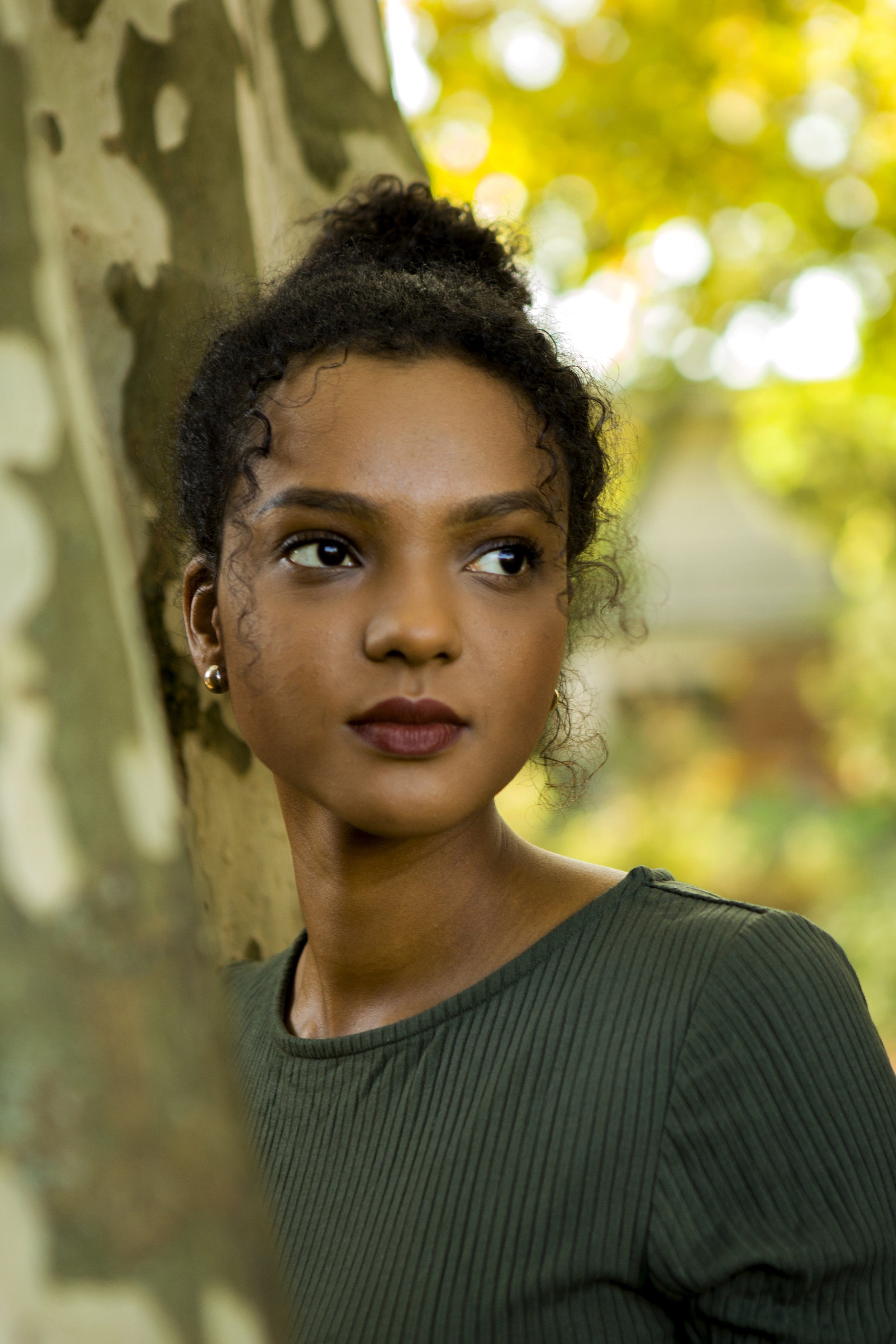 Nahaufnahme eines jungen afrikanischen Mädchens, das neben einem Baum steht und nach rechts schaut, im Hintergrund sind orange und gelbe Blätter, die auf einen herbstlichen Tag hinweisen.