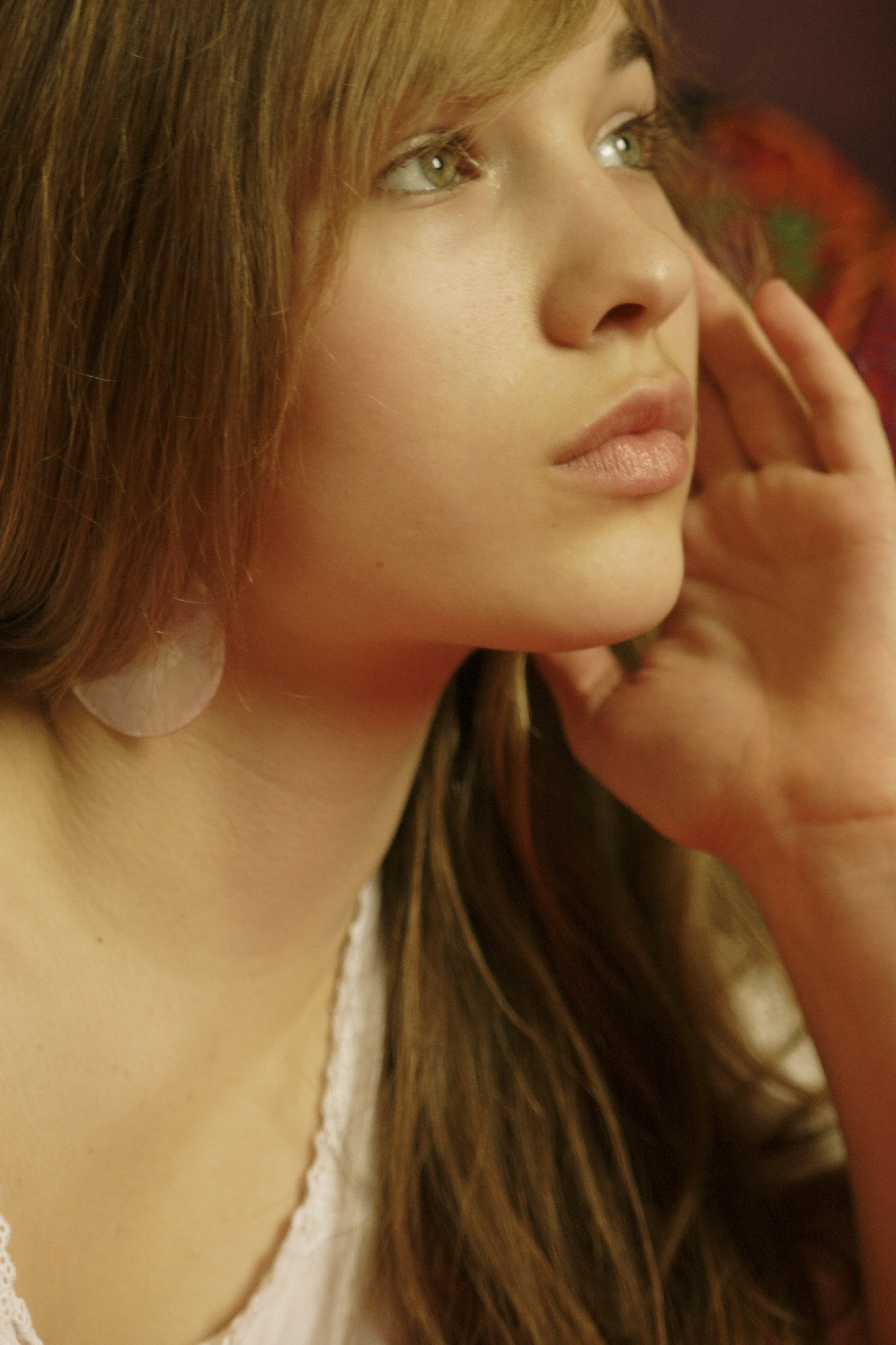 Nahaufnahme eines jungen Mädchens mit braunem, langem Haar, grünen Augen und natürlichem Make-up, das entspannt an einem Ohr berührt.