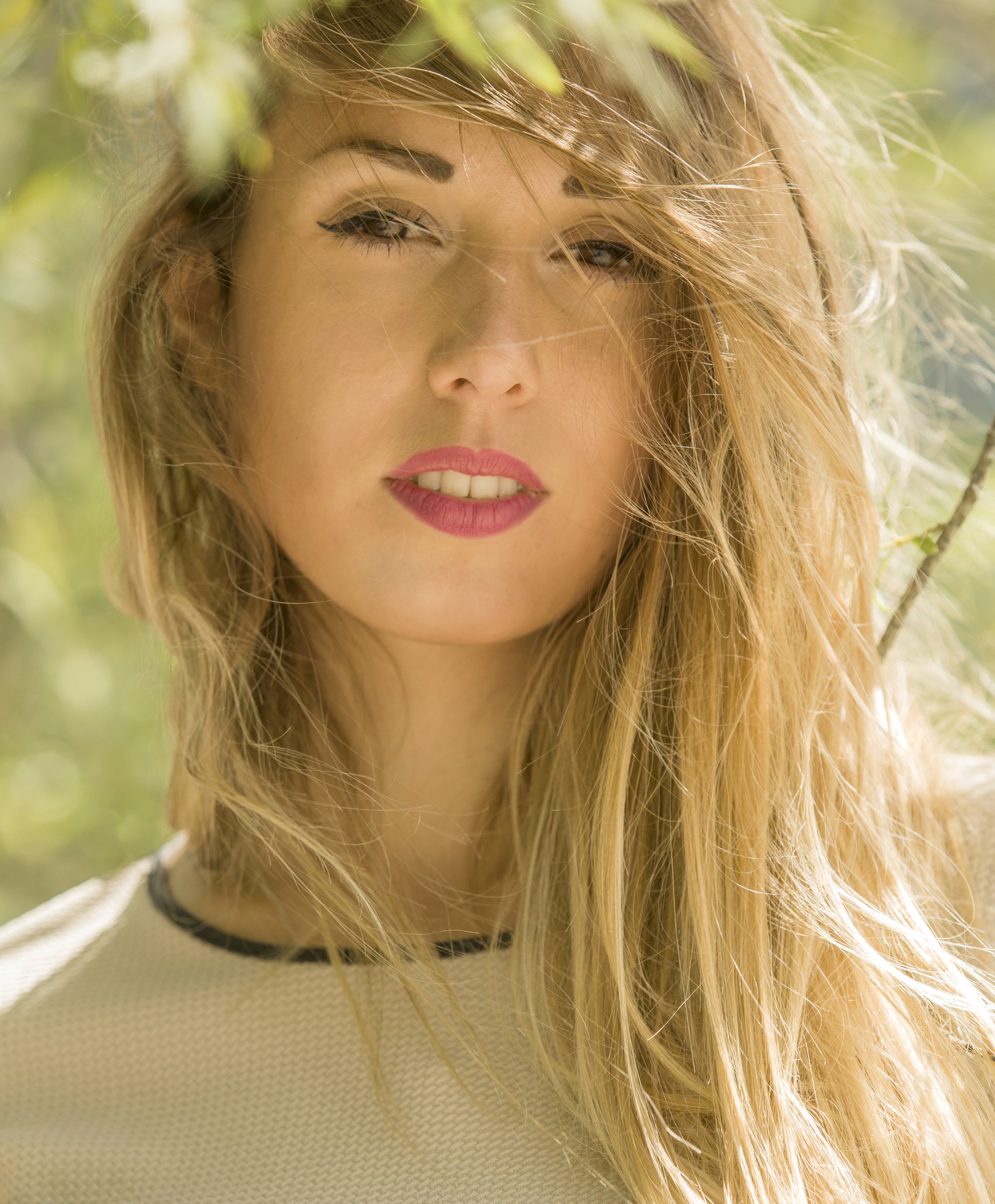 Nahaufnahme eines jungen Mädchens mit langen, lockigen, blonden Haaren, grünen Augen und Make-up, in natürlichem Licht mit unscharfem grünen Hintergrund.