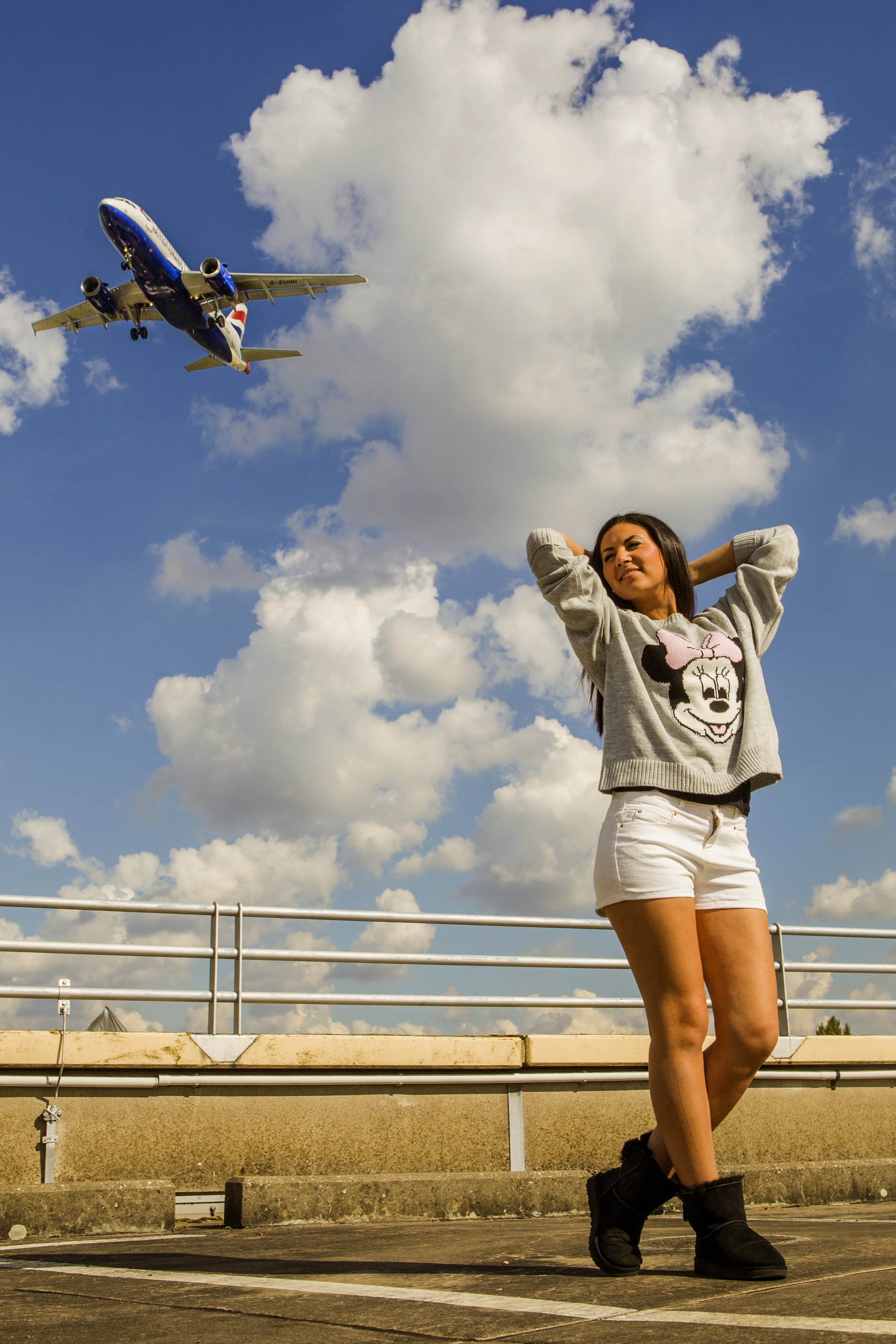 Eine junge Frau steht auf einem Parkplatz, trägt ein graues Sweatshirt mit Minnie Mouse und weiße Shorts, hat die Arme hinter dem Kopf und blickt in die Ferne. Ein Flugzeug fliegt am blauen Himmel mit Wolken vorbei.