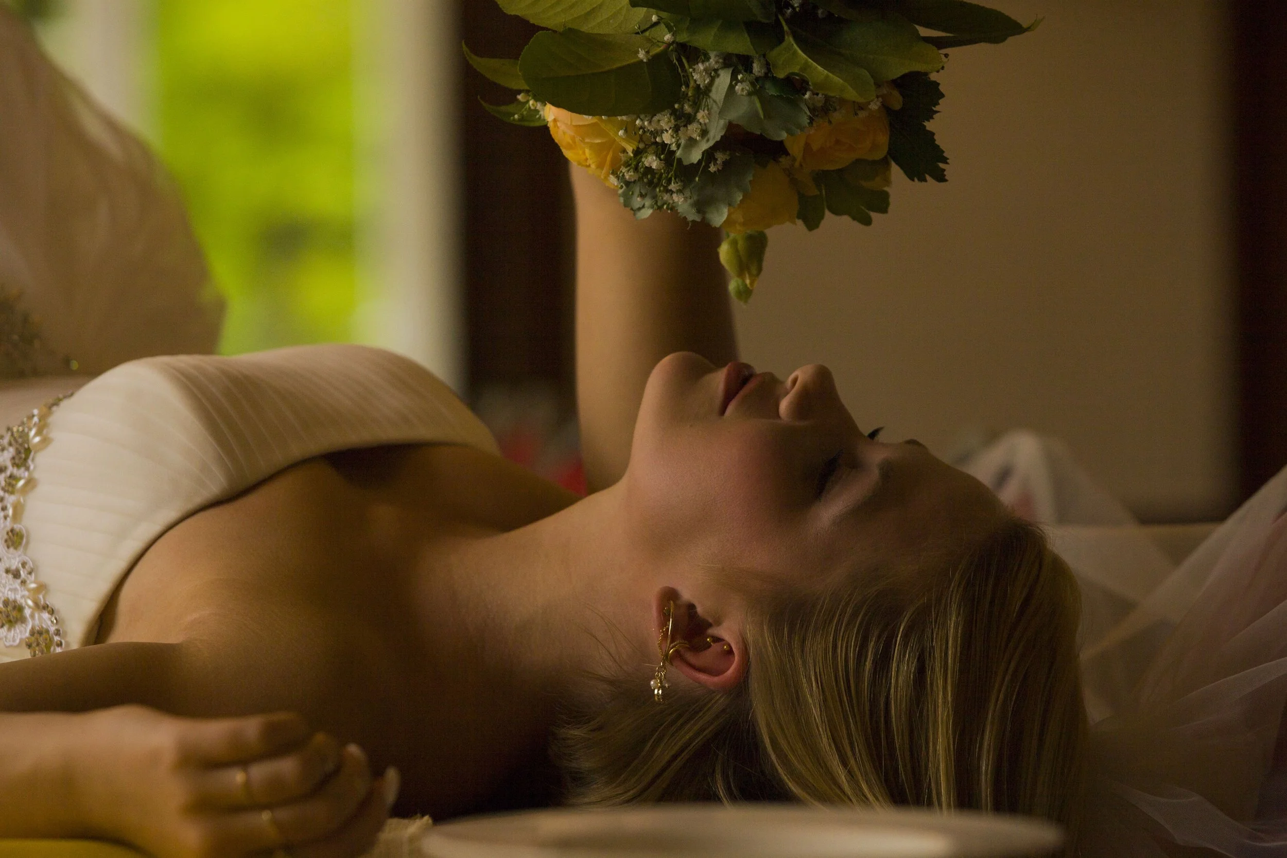 Eine Frau liegt auf einer Seite im Bett, hält einen Blumenstrauß mit gelben und weißen Blumen, hat ihre Augen geschlossen, trägt elegante Kleidung und goldene Ohrringe.