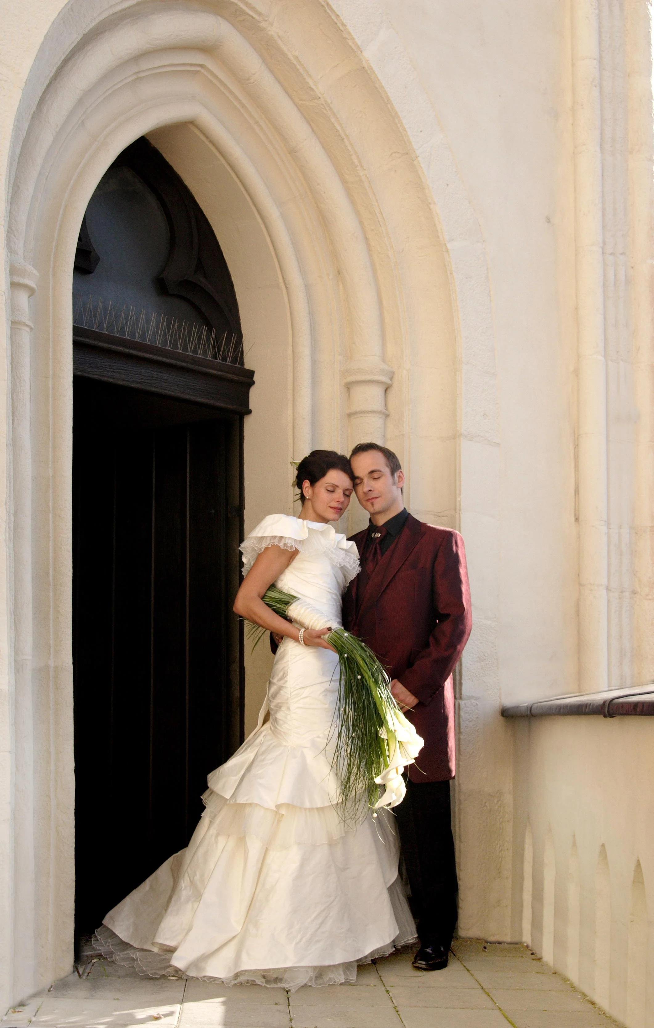 Ein Hochzeits-Paar steht vor einer alte Steinarchitektur, die Braut in einem weißen Kleid mit einem langen Rock hält einen Blumenstrauß, der Bräutigam trägt einen burgunderfarbenen Anzug.