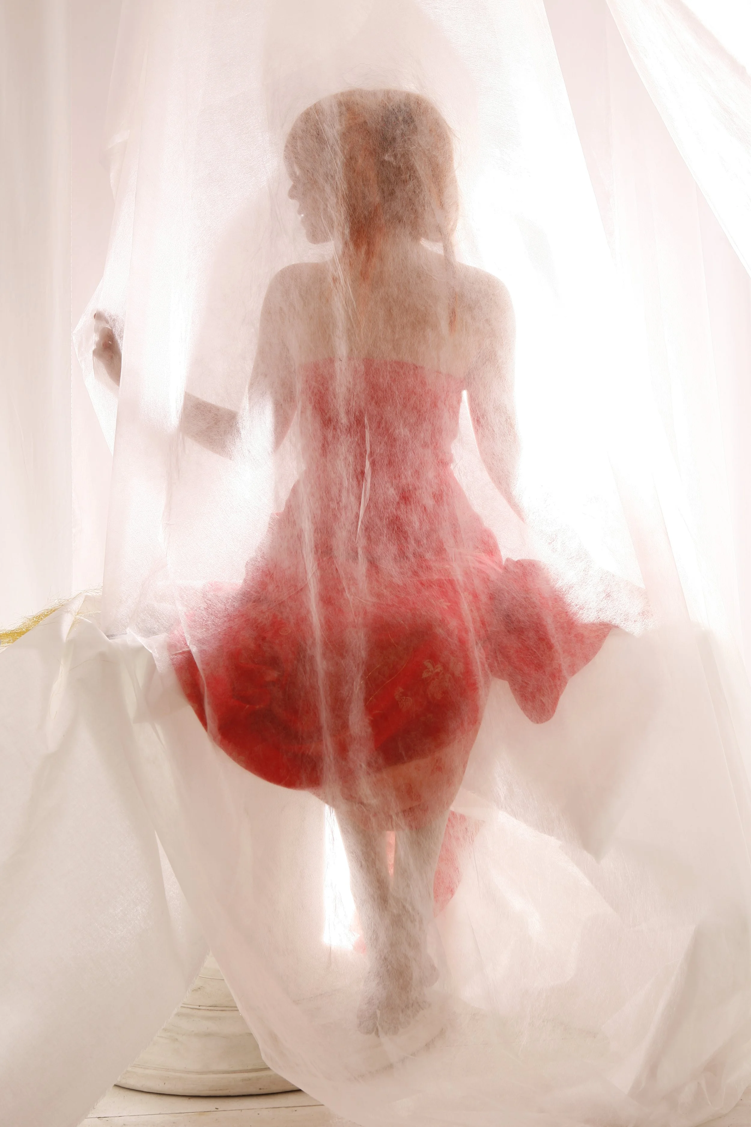 Eine Frau in einem roten Kleid sitzt hinter einem weißen, semi-transparenten Tuch, das sie teilweise verdeckt.