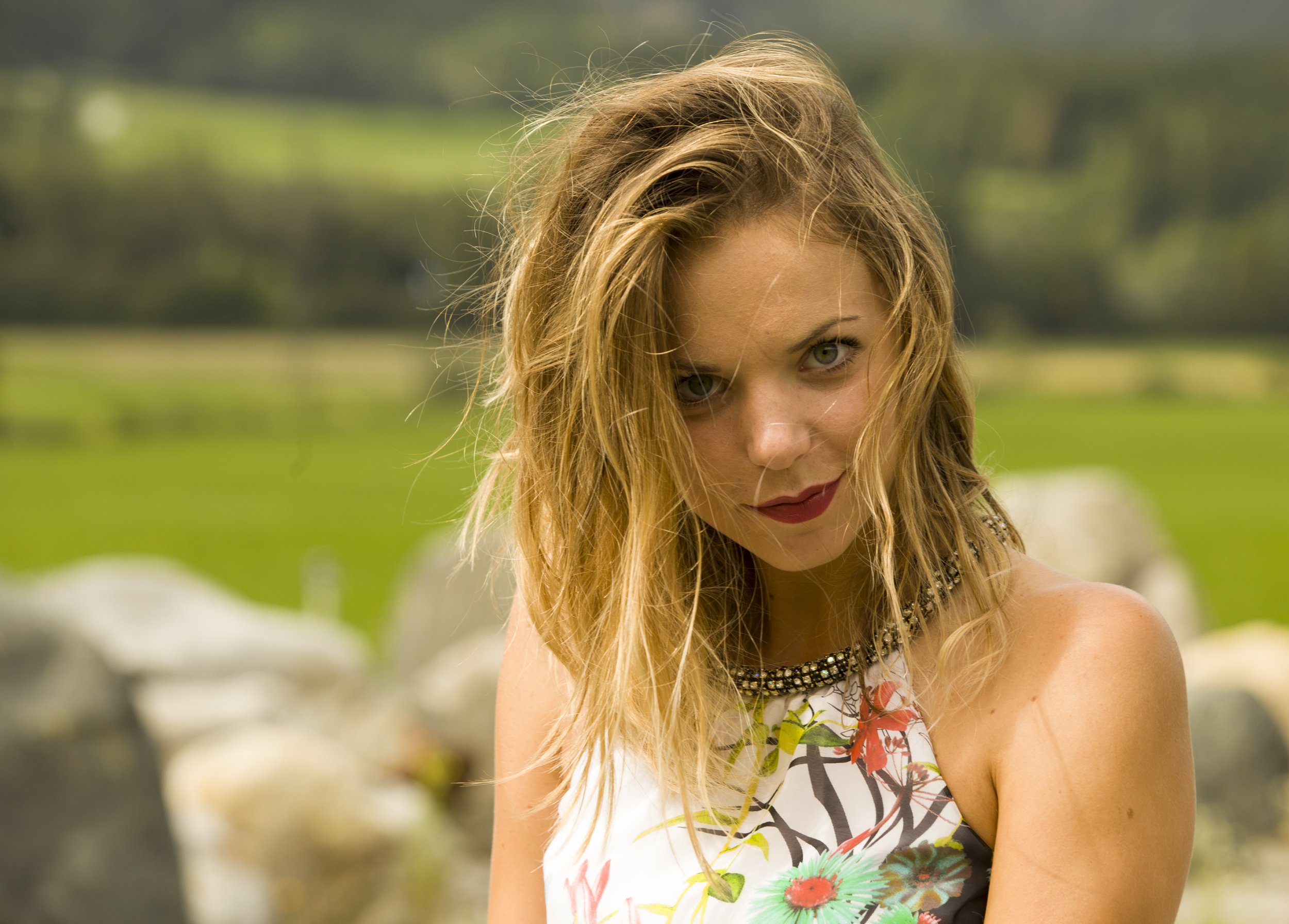 Junge Frau mit blonden, gewellten Haaren, trägt ein sommerliches Kleid mit Blumenmuster, steht im Freien vor grüner Natur.
