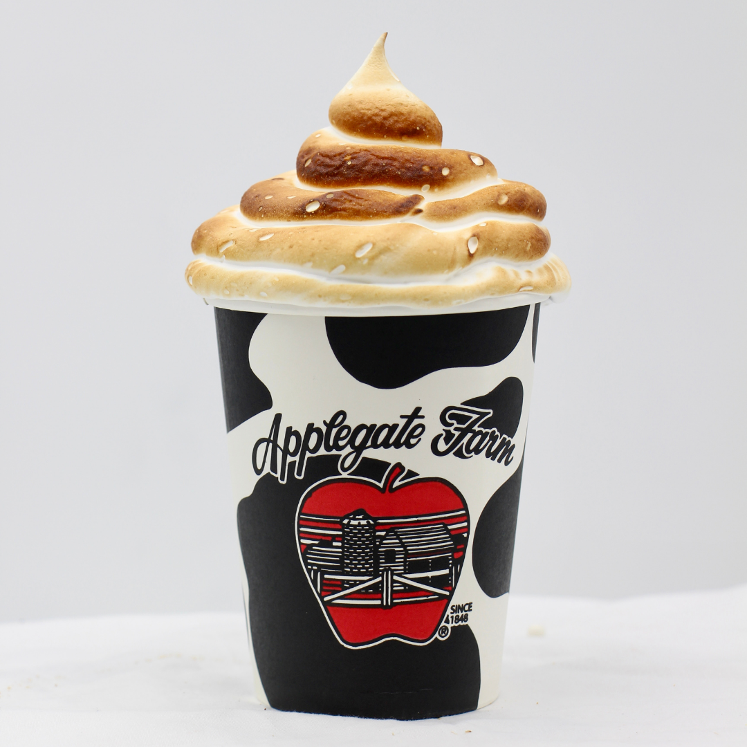 A paper cup with black and white cow print pattern, featuring the text "Applegate Farm" and a red apple with a farm scene illustration. The cup is topped with toasted meringue.