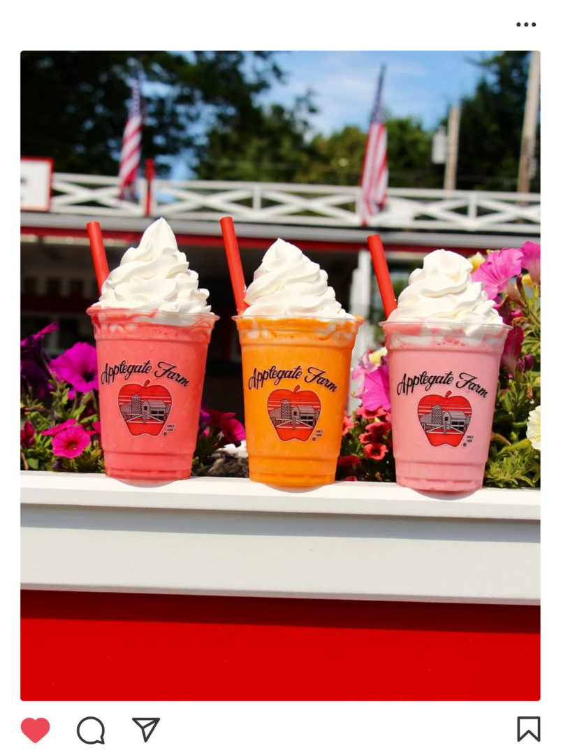 Three colorful smoothies topped with whipped cream, in cups labeled 'Applegate Farm,' placed in a flower box with pink and white flowers, outdoors with American flags in the background.