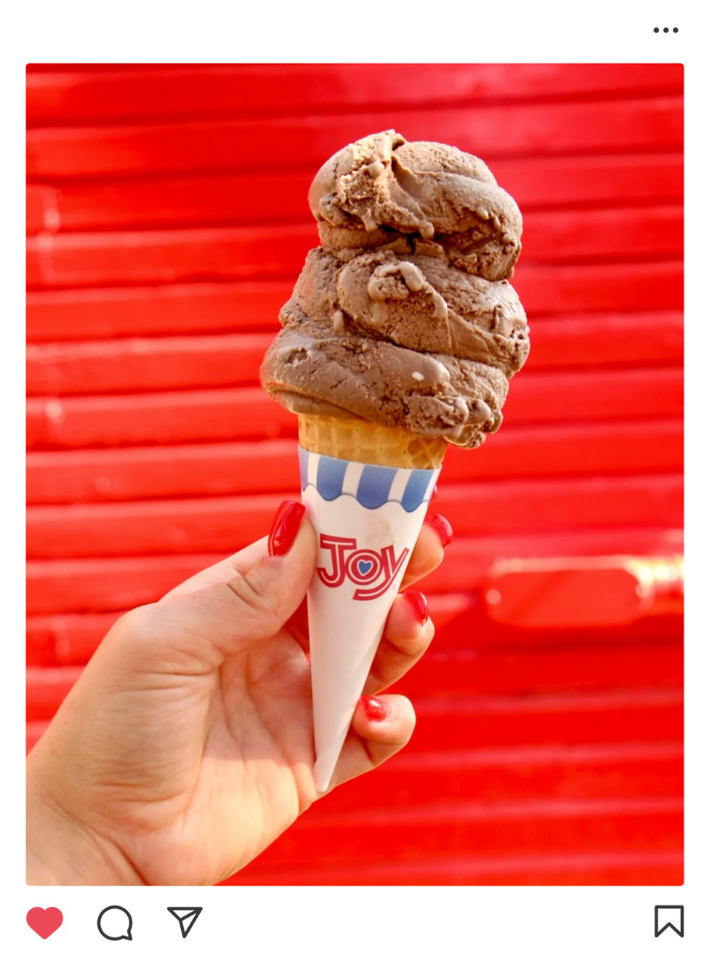 A hand with red-painted nails holding a chocolate ice cream cone against a red wooden background.