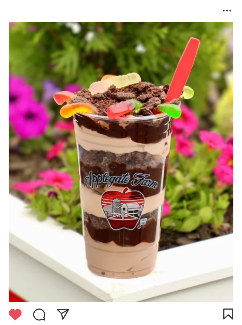 Chocolate and vanilla layered ice cream sundae in a clear cup with green and red gummy candies, cookies, and a red spoon, labeled 'Applegates Farm', outdoors with pink flowers in the background.