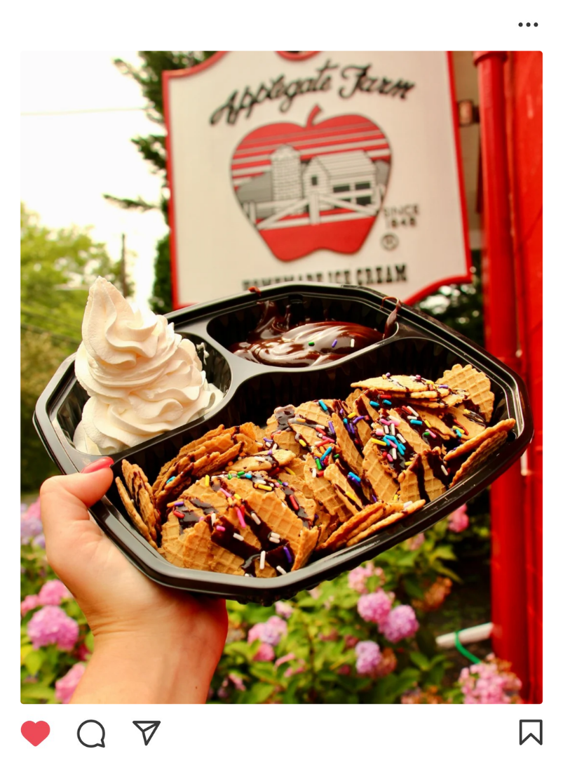 A person holding a black plastic tray with vanilla swirl soft serve ice cream, chocolate sauce with sprinkles, and a row of waffle cookies drizzled with chocolate and decorated with colorful sprinkles, in front of a red and white apple-shaped sign for Appalachian Farm.