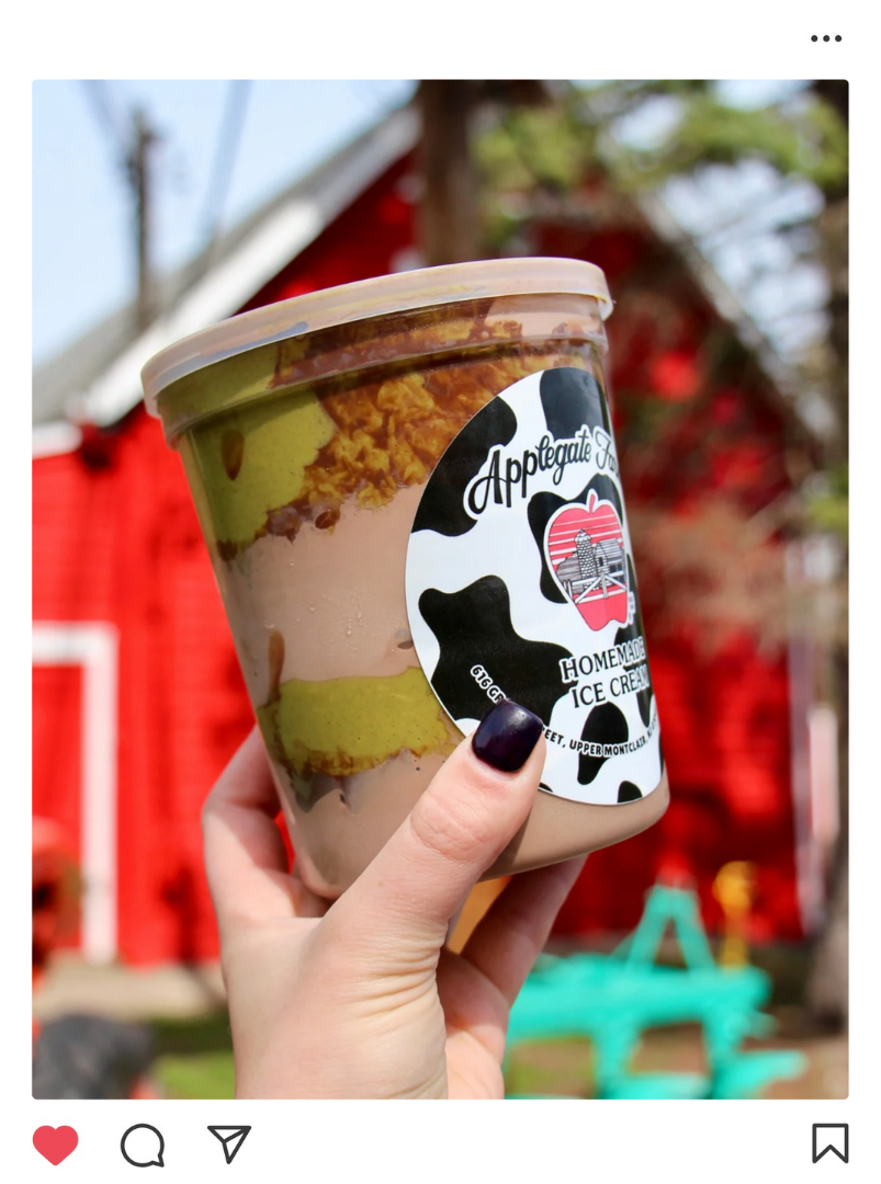 Hand holding a cup of homemade ice cream with chocolate, vanilla, and green layers. The cup has a black and white cow print design and the label reads 'Applegate Farm Homemade Ice Cream.' In the background, there is a red barn and trees.