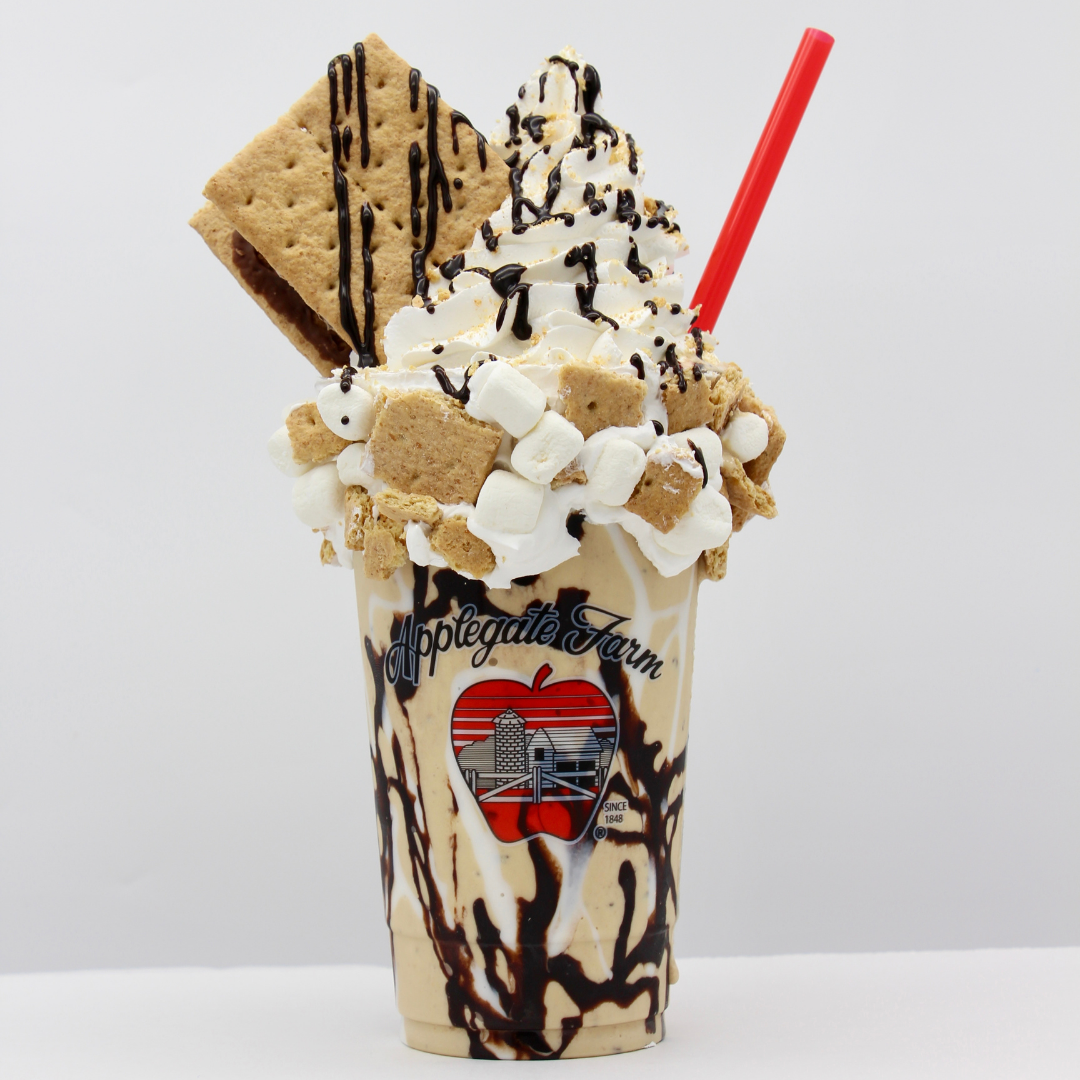 A milkshake with whipped cream, chocolate syrup, graham crackers, mini marshmallows, and a large cookie, in a glass with 'Applegate Farm' logo and a red straw.