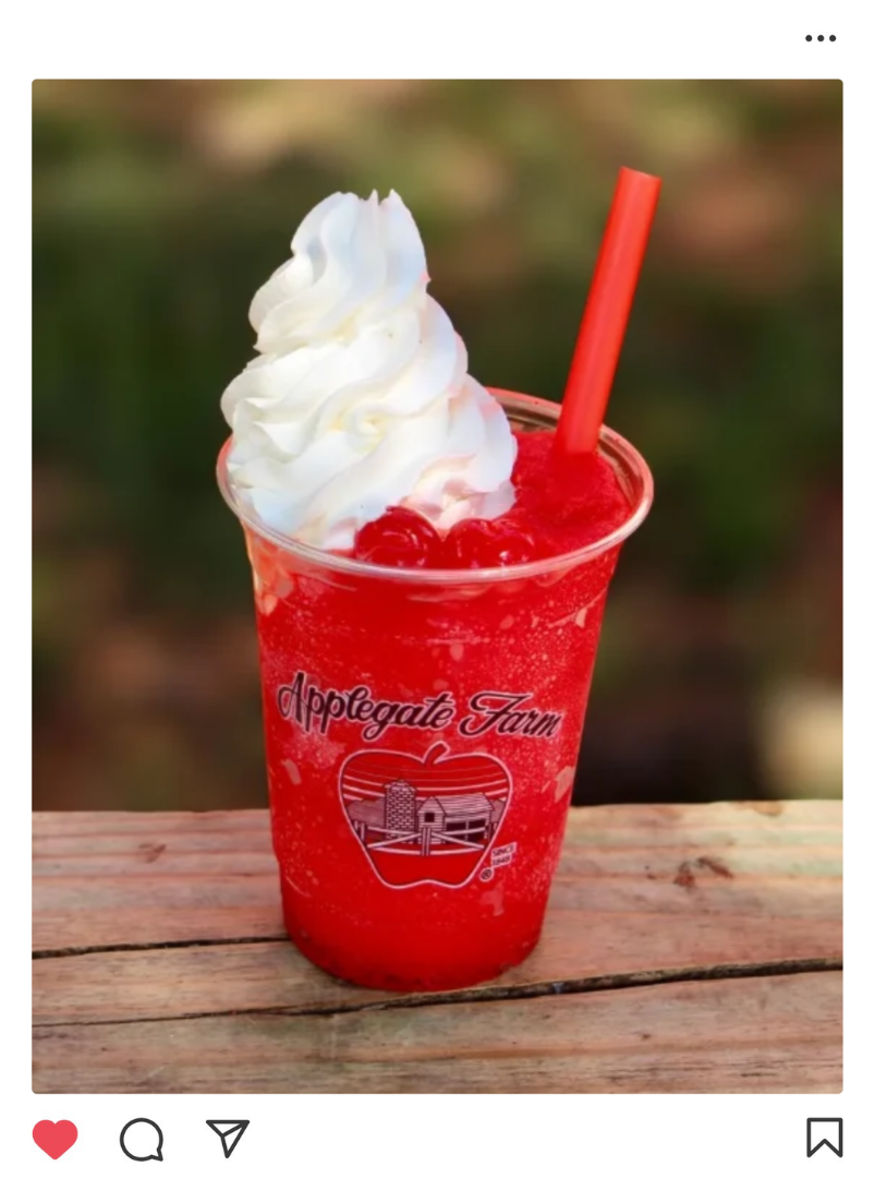 A red strawberry slush drink with whipped cream on top, served in a clear plastic cup with a red straw. The cup has a logo that says 'Applegate Farm' with a barn and apple illustration, placed on a wooden surface.