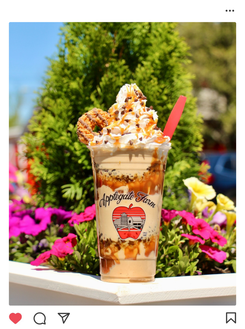 A large milkshake with whipped cream, caramel drizzle, chocolate sprinkles, and a cookie, in a clear plastic cup with Applegate Farm logo, surrounded by flowers.