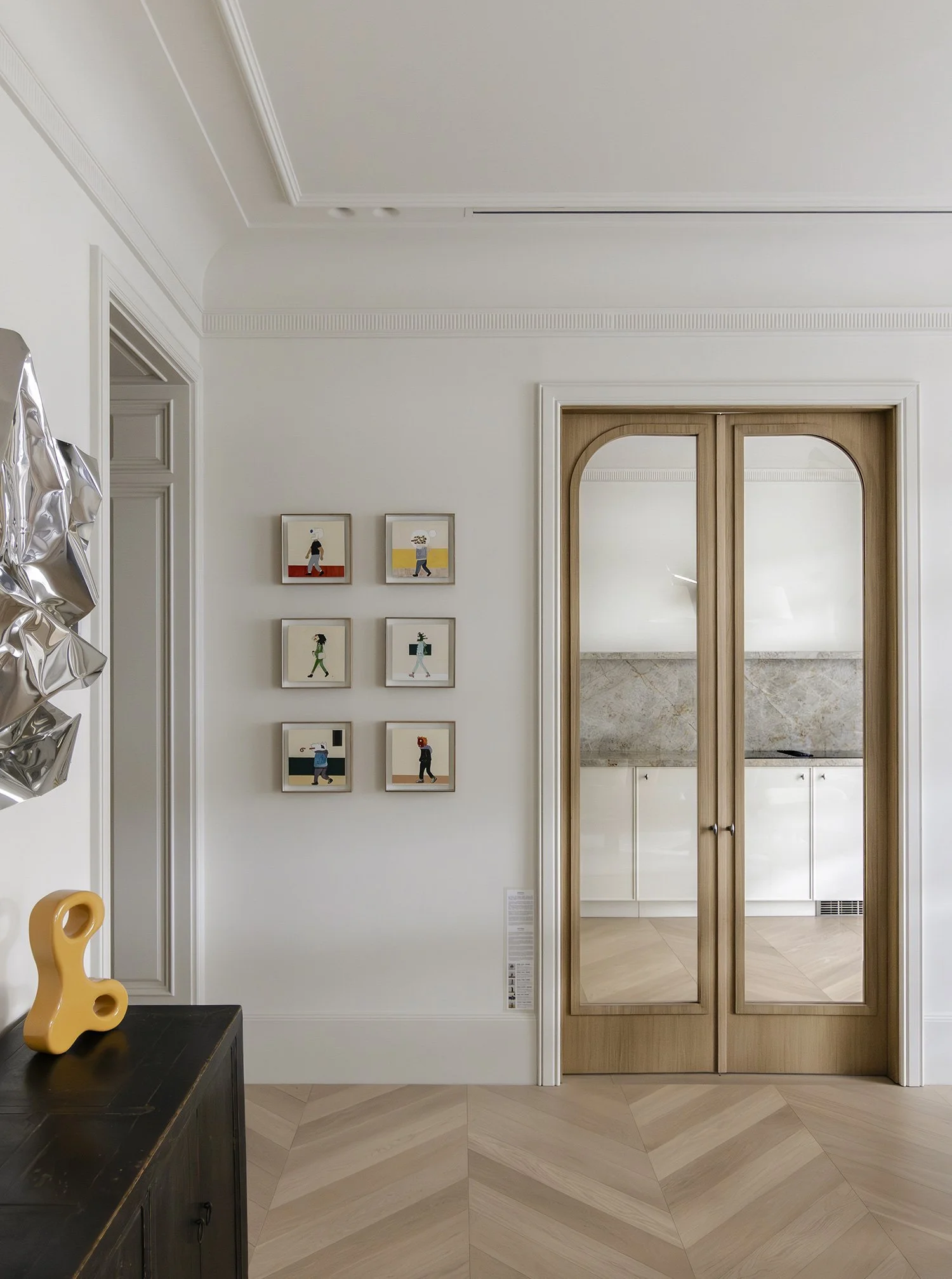 Sala de estar con paredes blancas, ocho cuadros decorativos con figuras humanas en diferentes poses, puertas de madera con reflejo en las ventanas, suelo de madera en zigzag, y una mesa negra con una escultura amarilla en forma de curva.