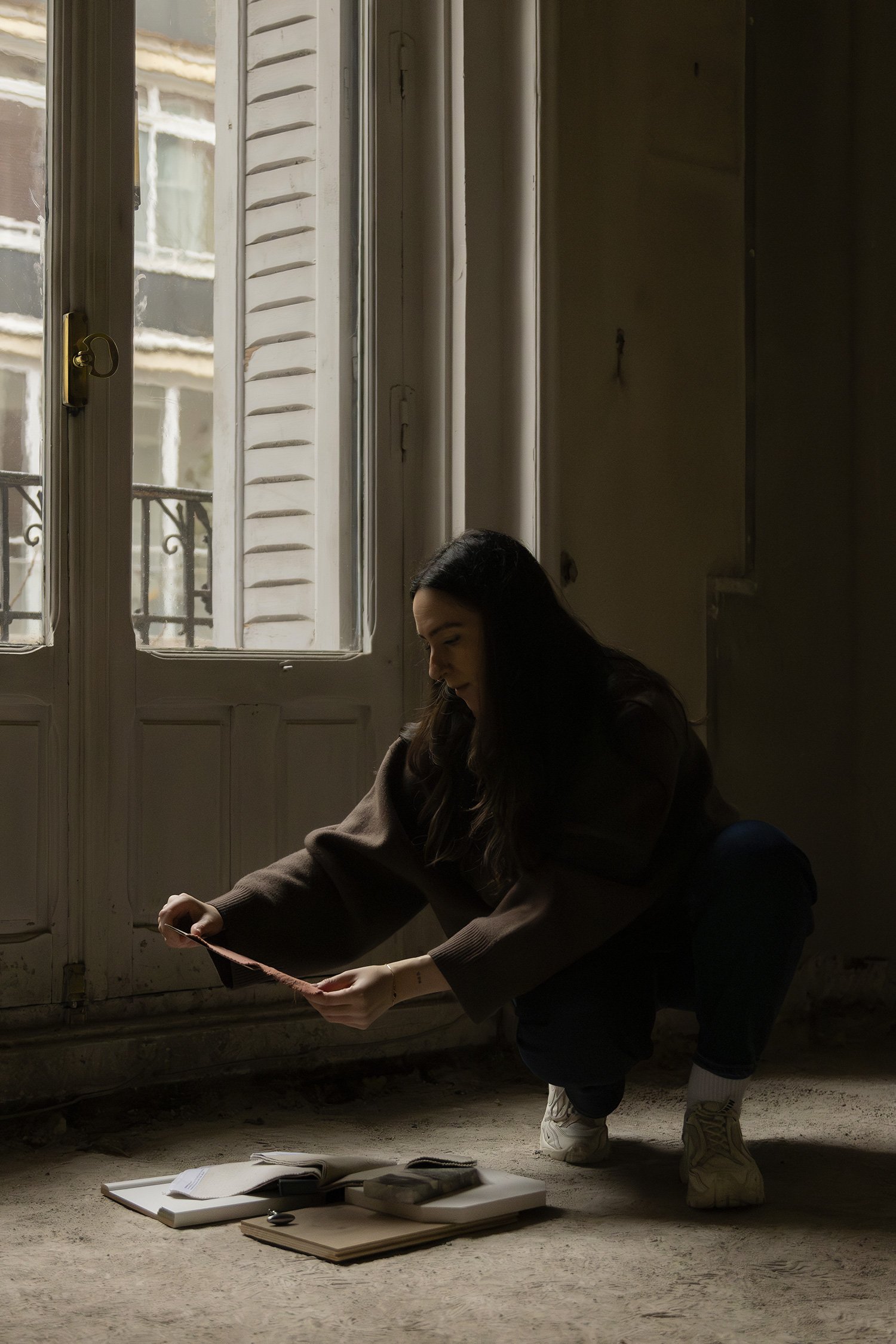 Una mujer con cabello largo y oscuro, vestida con un suéter marrón y pantalones oscuros, se agacha en un suelo cubierto de polvo o arena, sosteniendo una regla o cinta métrica. Frente a ella hay varias carpetas o libretas apiladas y algunos objetos sobre ellas. La escena está en una habitación con paredes de color beige y ventanas grandes con persianas blancas, que dejan entrar luz natural.