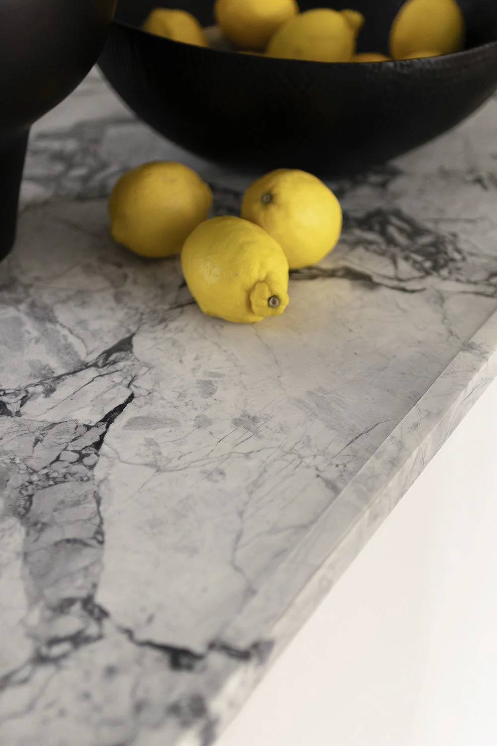 Cuatro limones en una superficie de mármol con un recipiente negro de frutas al fondo.