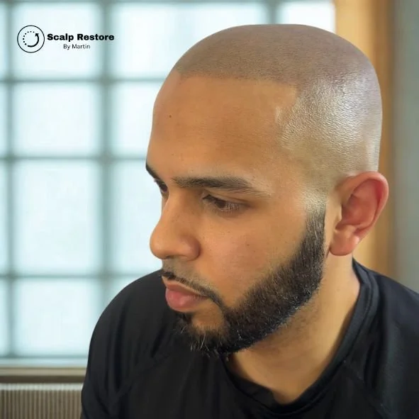 Close-up of a man with a shaved head and beard, sitting indoors in front of a window.