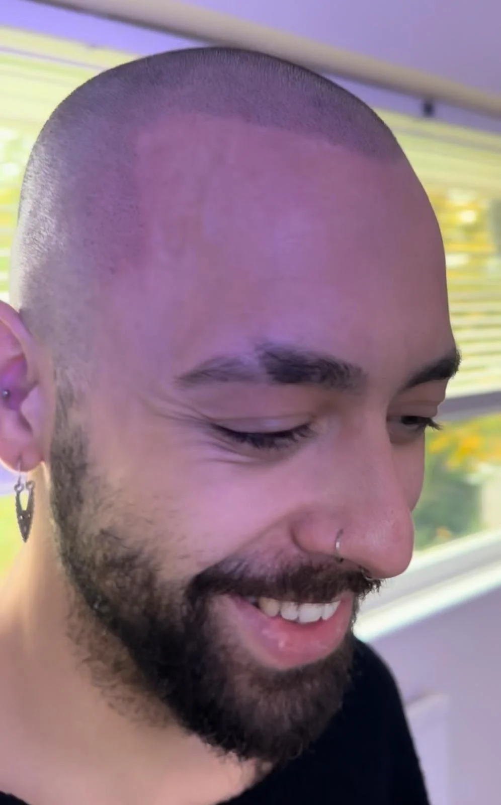 Close-up of a smiling man with a shaved head, beard, and multiple earrings, looking downward, with a window and yellow-striped background behind him.