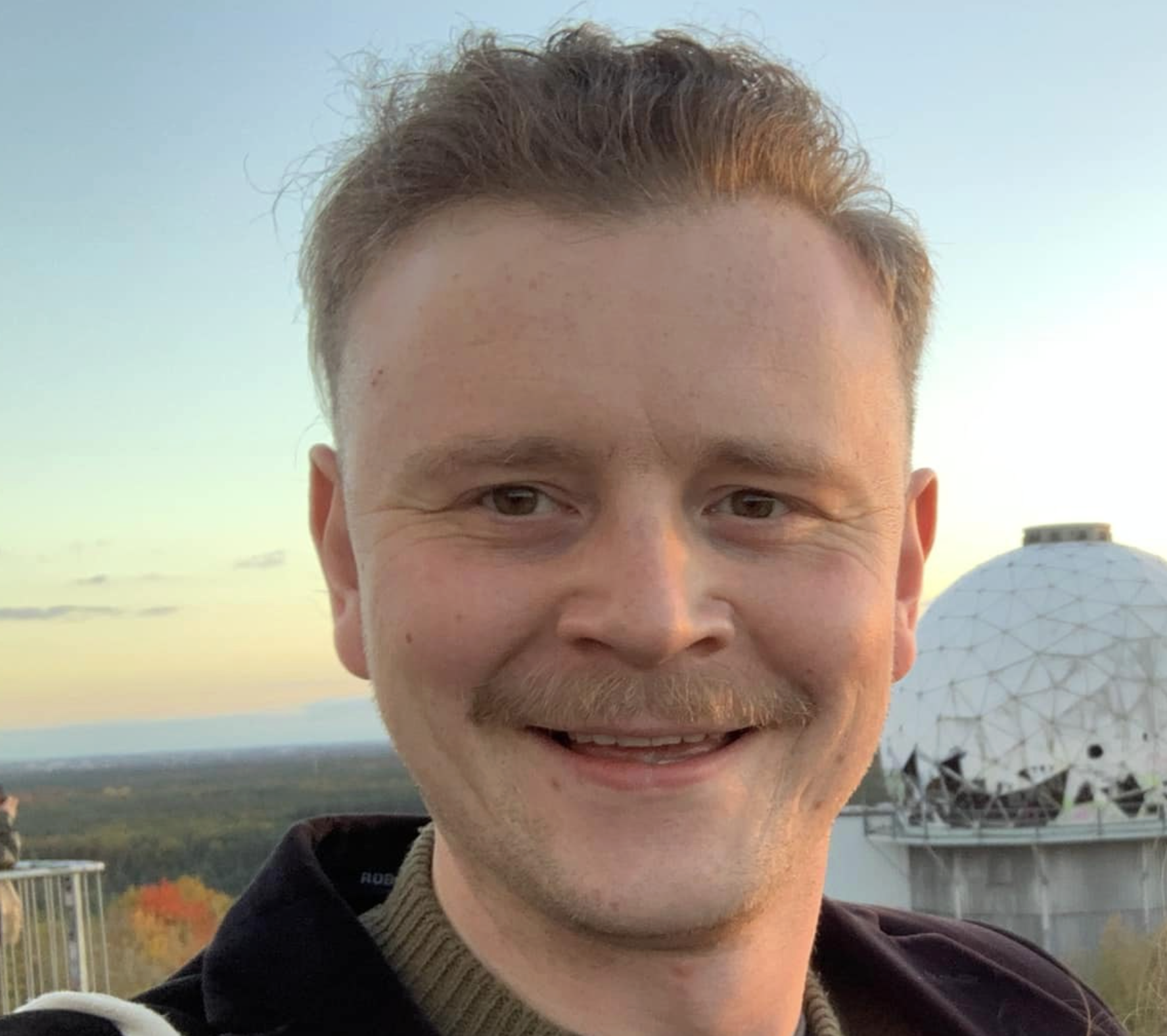A smiling man with short, light brown hair and a goatee, taken outdoors during sunset. In the background, a large white spherical observatory dome is visible against a landscape with distant trees and a sky with soft clouds.