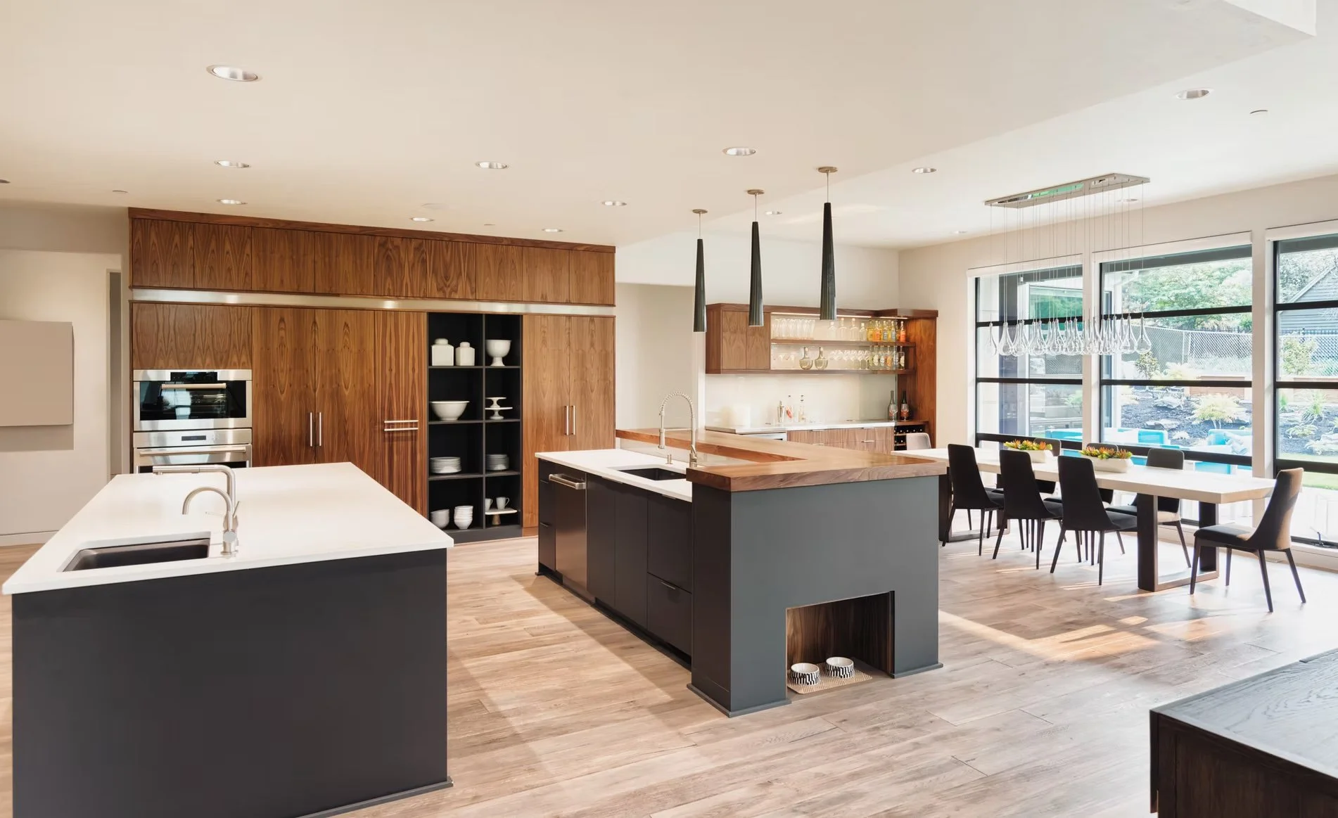 Cocina moderna con isla negra y paredes de madera, con área de comedor con sillas negras junto a grandes ventanas que dejan entrar luz natural.