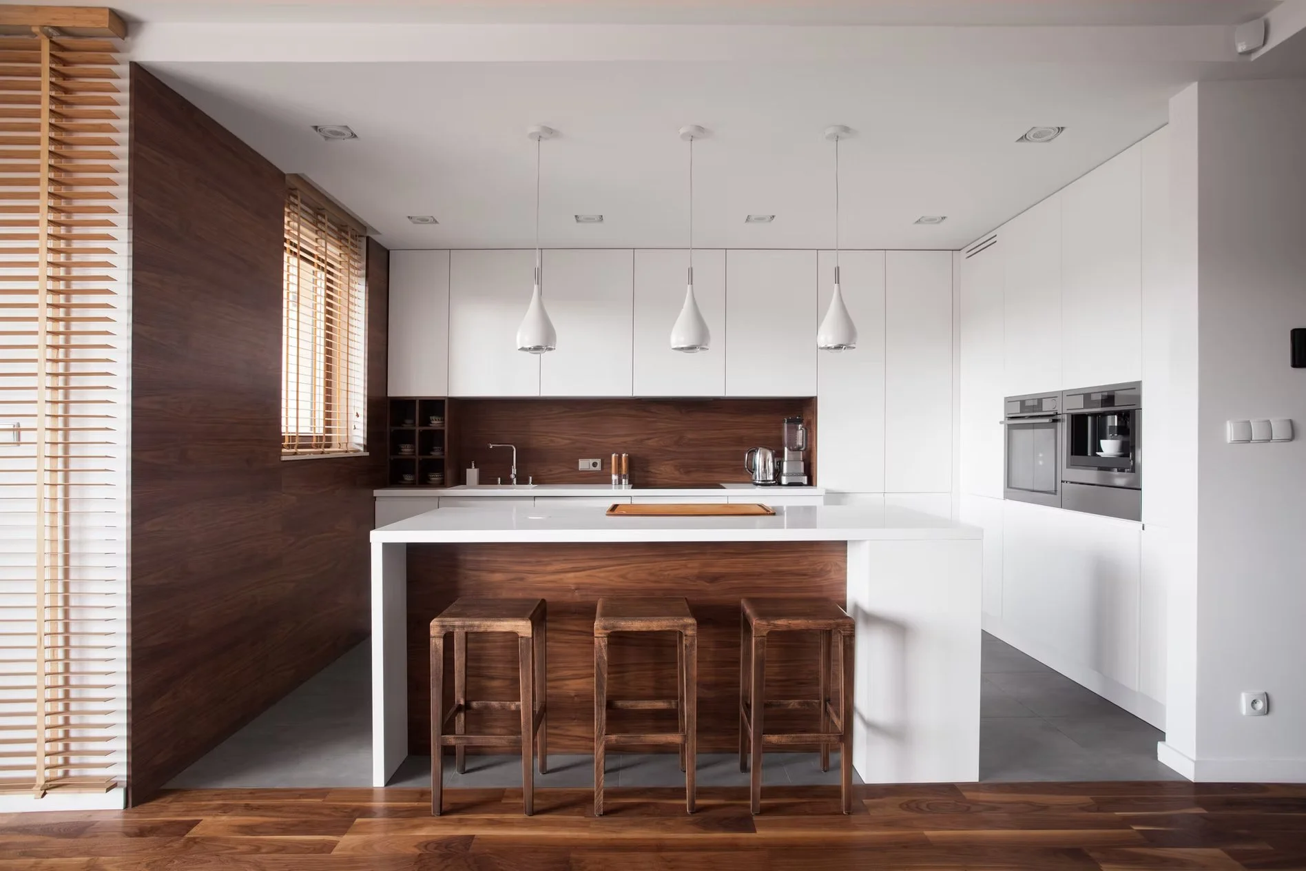 Cocina moderna con encimera blanca y paredes de madera, con tres taburetes de madera y ventanas con persianas de madera.