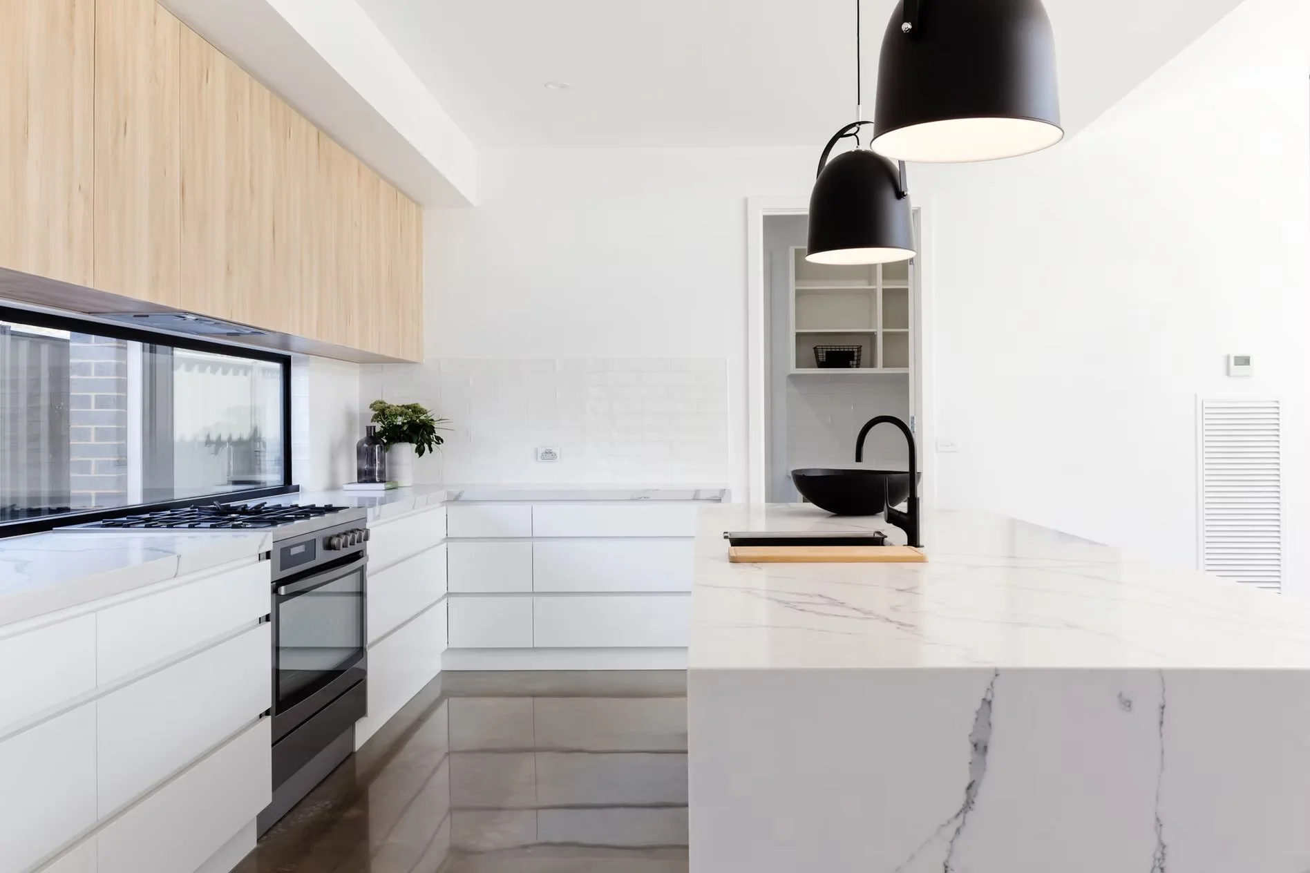 Cocina moderna con encimeras de mármol blanco, gabinetes blancos, estufa de acero, lámparas negras colgantes y ventanas grandes que permiten entrada de luz natural.