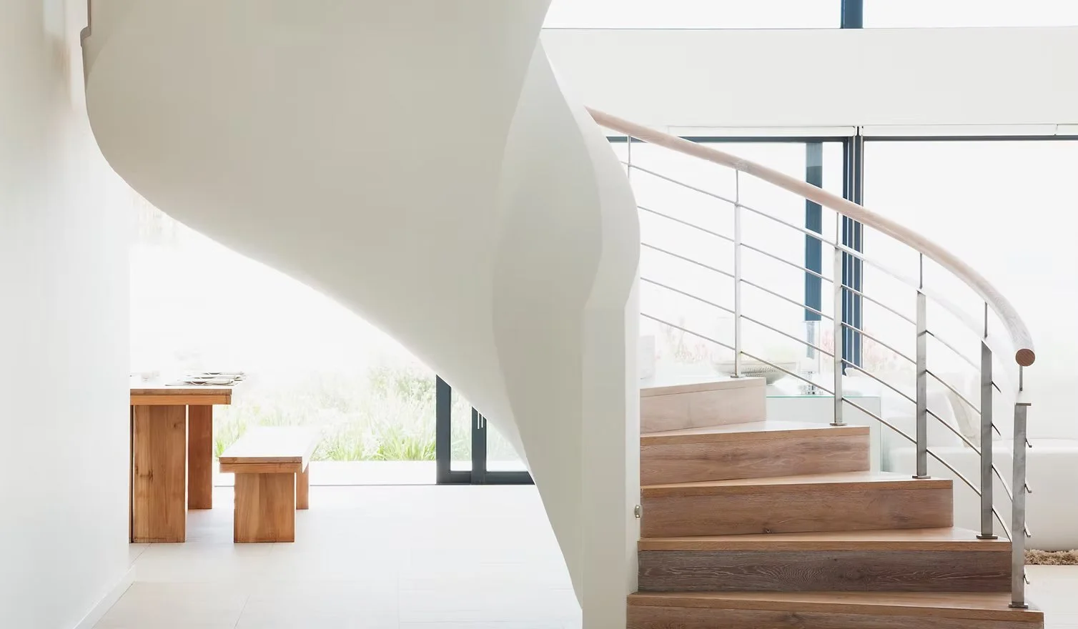Escalera de madera con barandales de metal en un interior luminoso y minimalista, con ventanas grandes al fondo.
