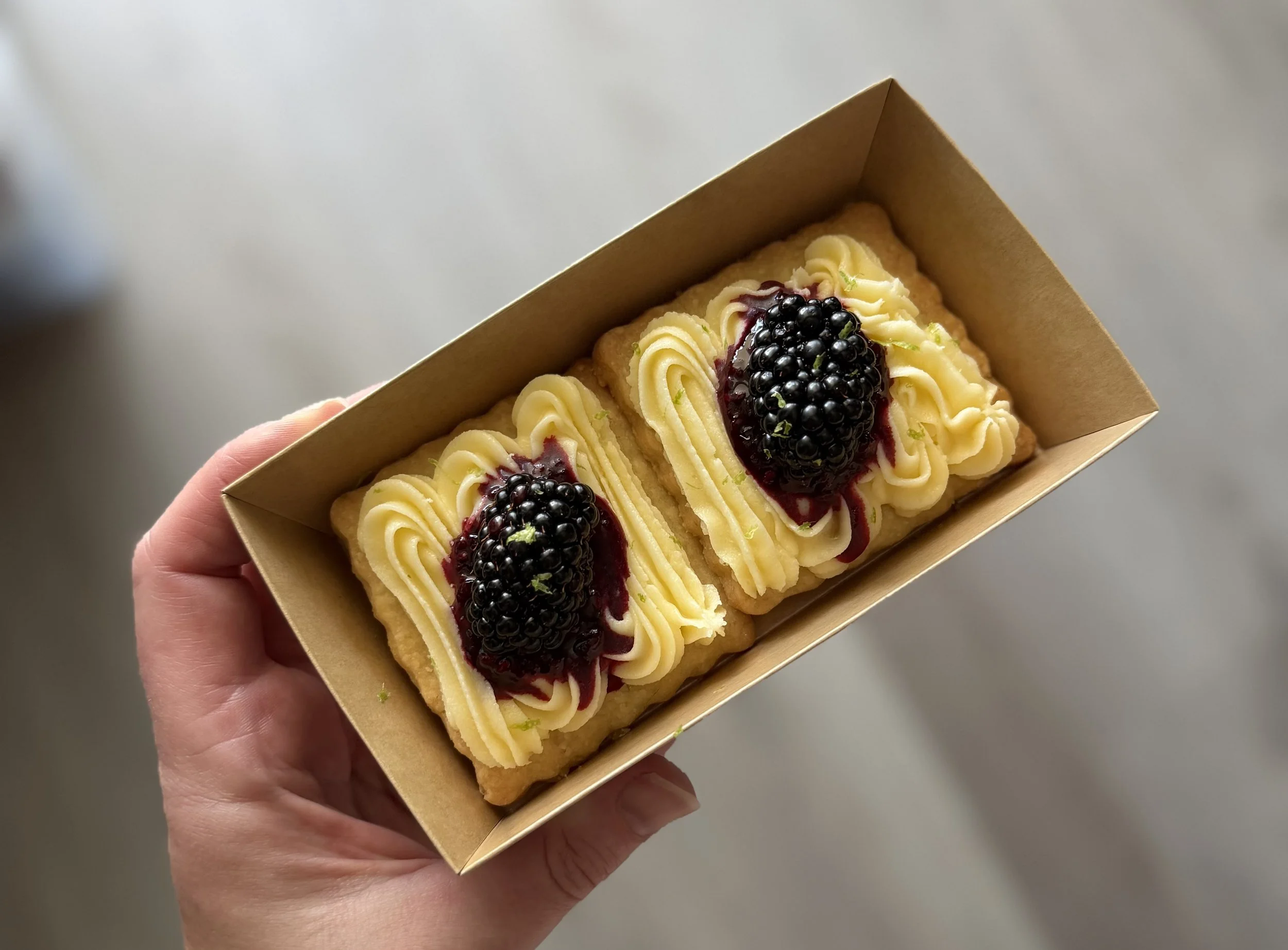 A hand holding a rectangular pastry topped with piped vanilla cream and blackberries, served in a small cardboard tray.