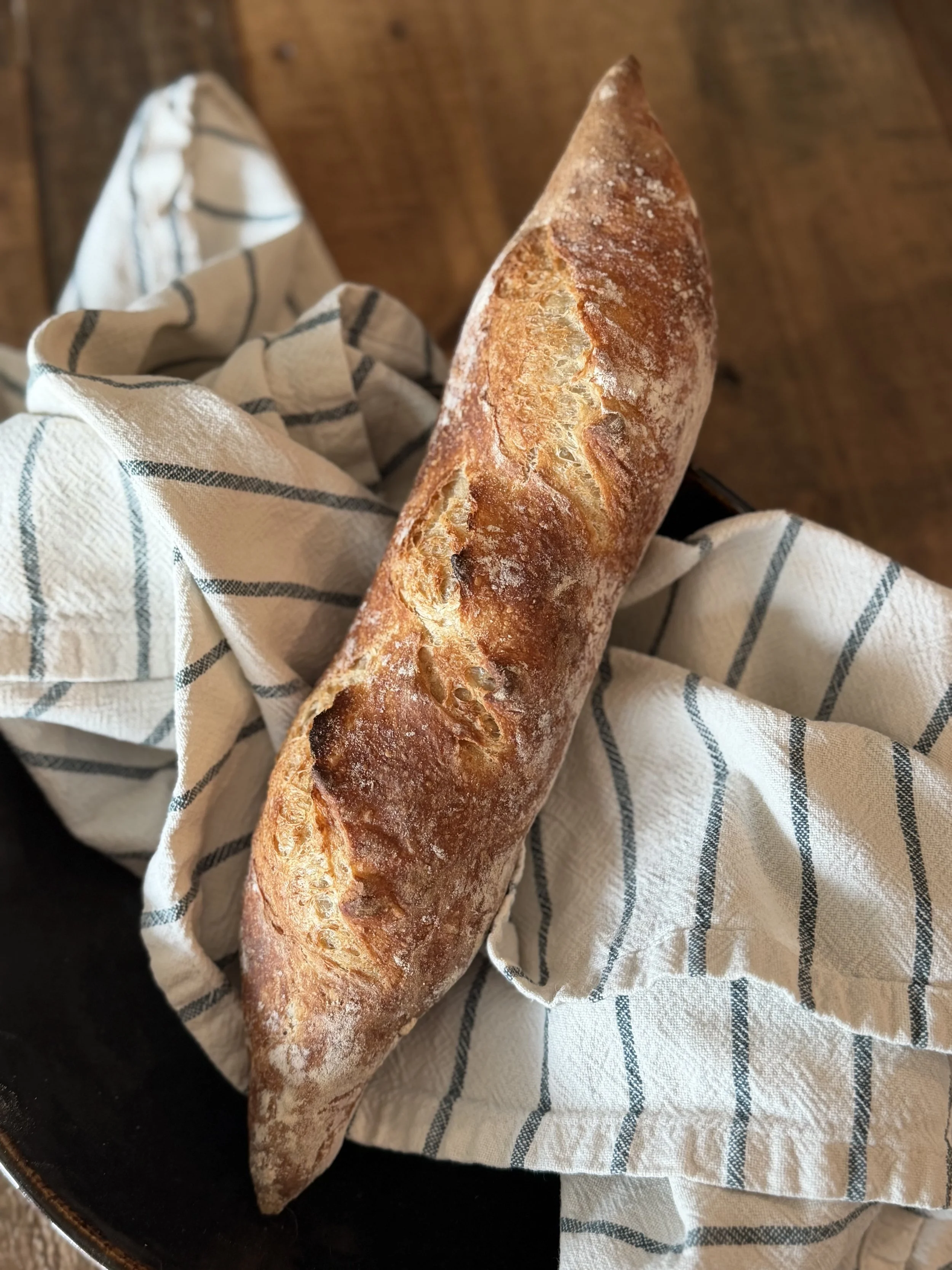 A freshly baked baguette with a golden brown crust resting on a white and blue striped cloth, placed in a black skillet.