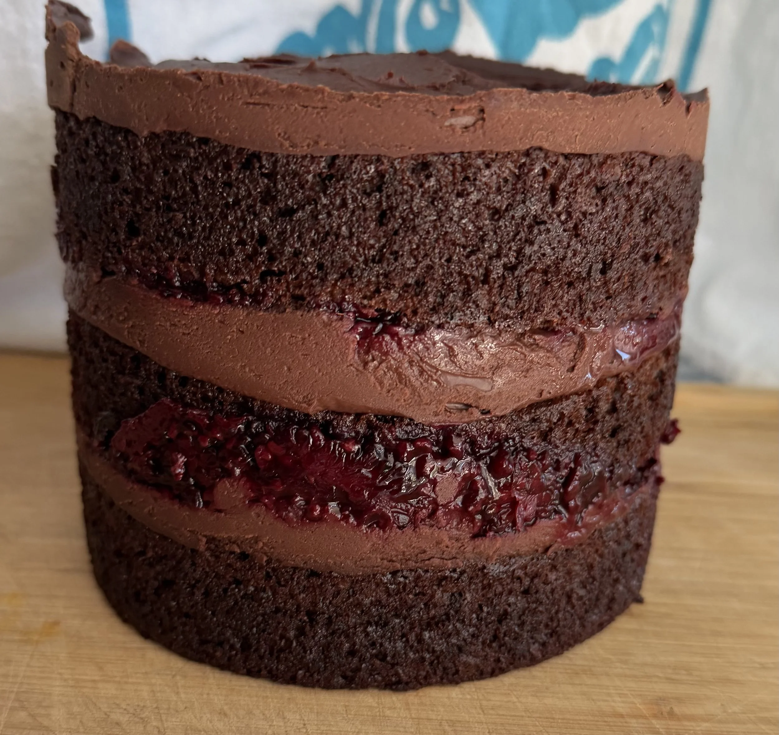 A close-up of a chocolate cake with three layers, featuring a smooth chocolate frosting on top and between the layers, with a textured dark cake at the bottom, placed on a wooden surface.