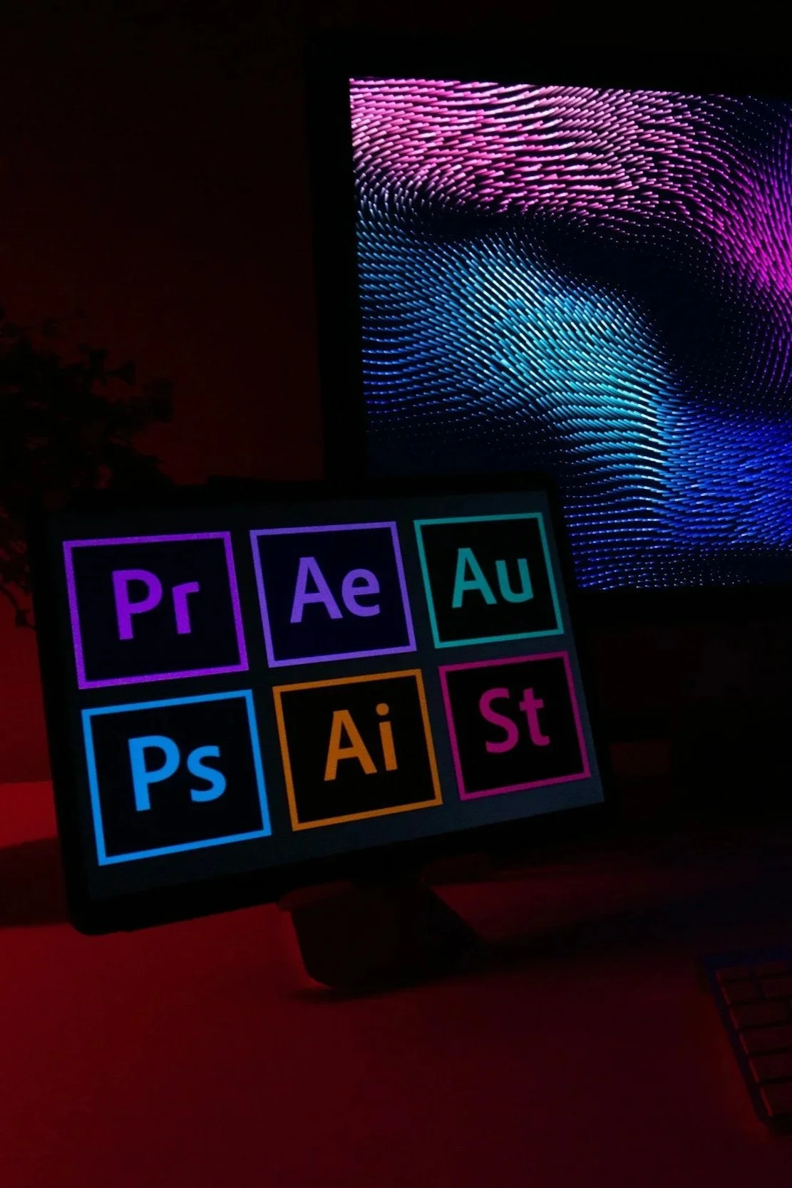 A computer monitor displays colorful images behind a graphic tablet showing icons of Adobe software programs, including Premiere Pro, After Effects, Audition, Photoshop, Illustrator, and Spark.