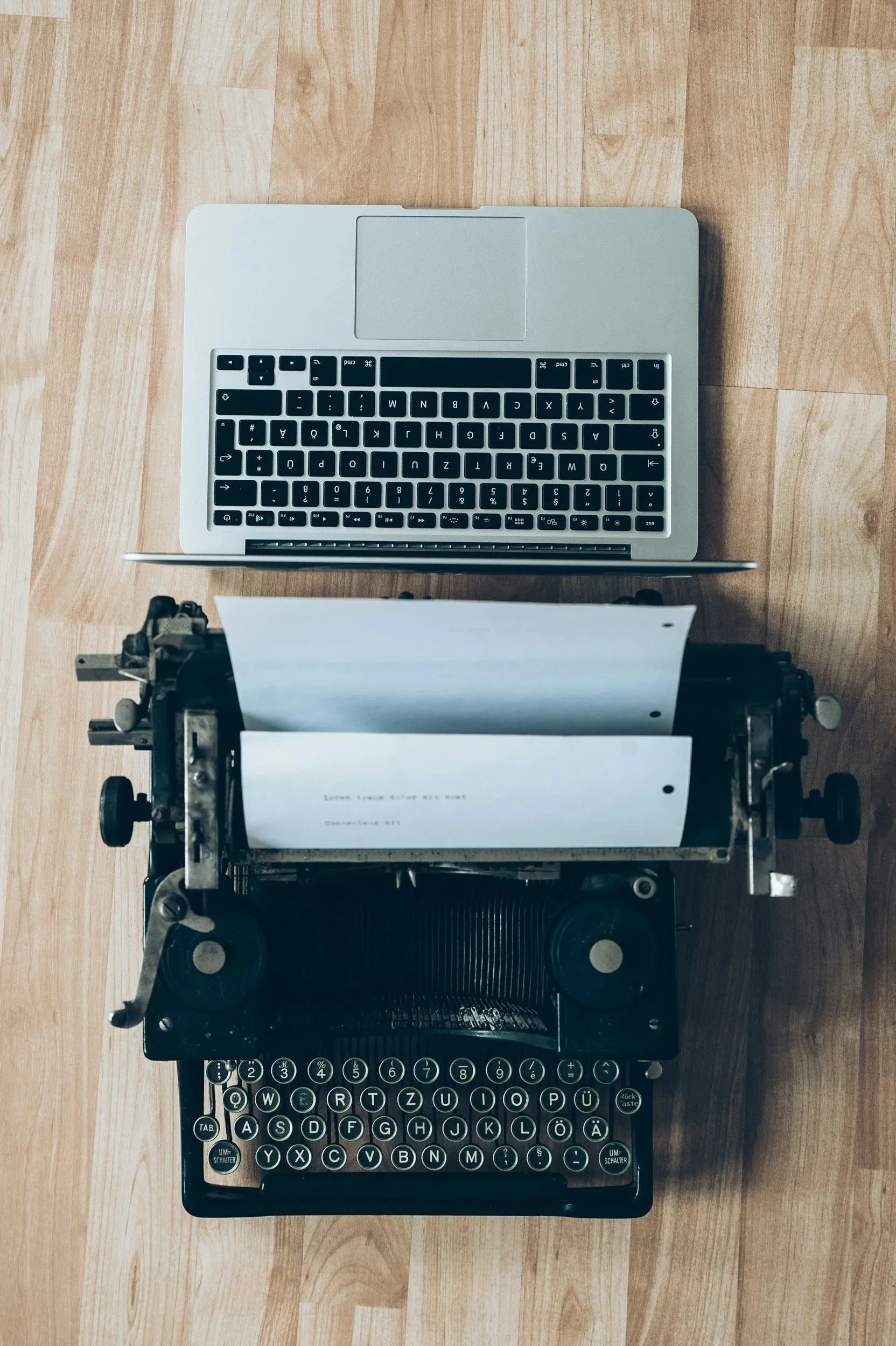 A vintage typewriter with paper loaded, positioned below a modern silver laptop on a wooden floor.