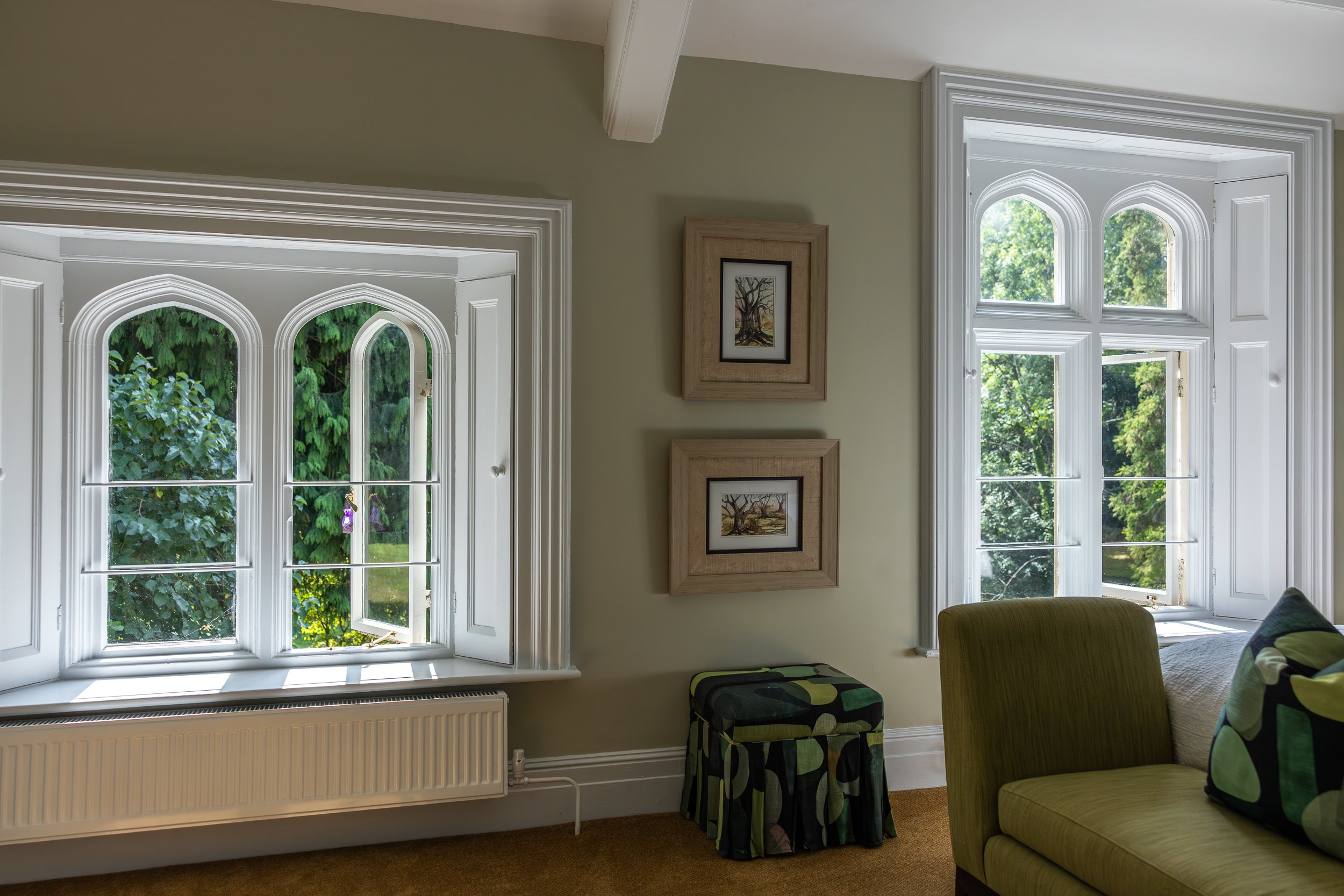Interior of a living room with two arched windows showing green trees outside, framed artwork on the wall, a patterned ottoman, and a green sofa with a cushion.