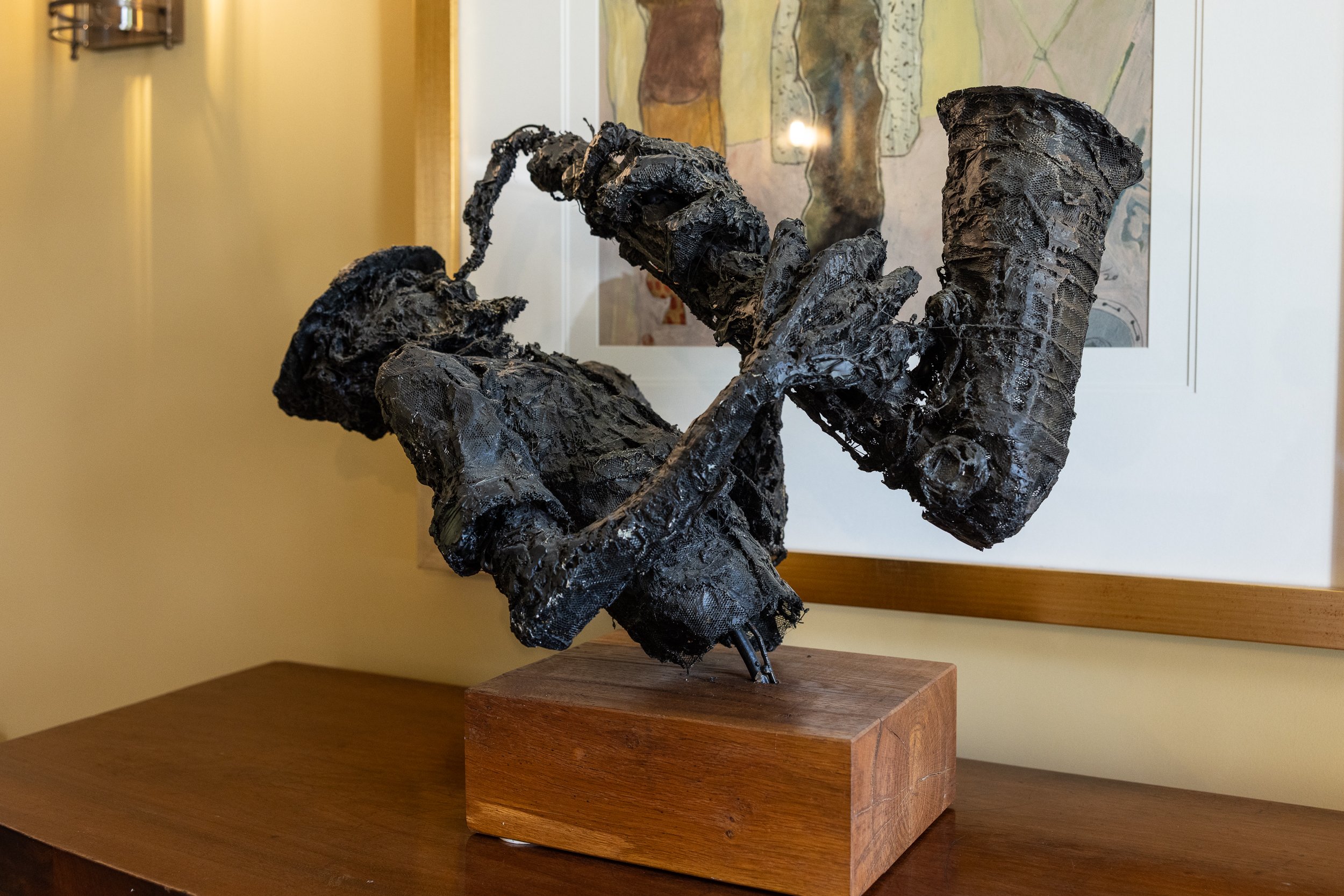 A sculpture of a person playing a violin, made with dark, textured material, mounted on a wooden base, displayed indoors against a wall with a framed artwork behind it.