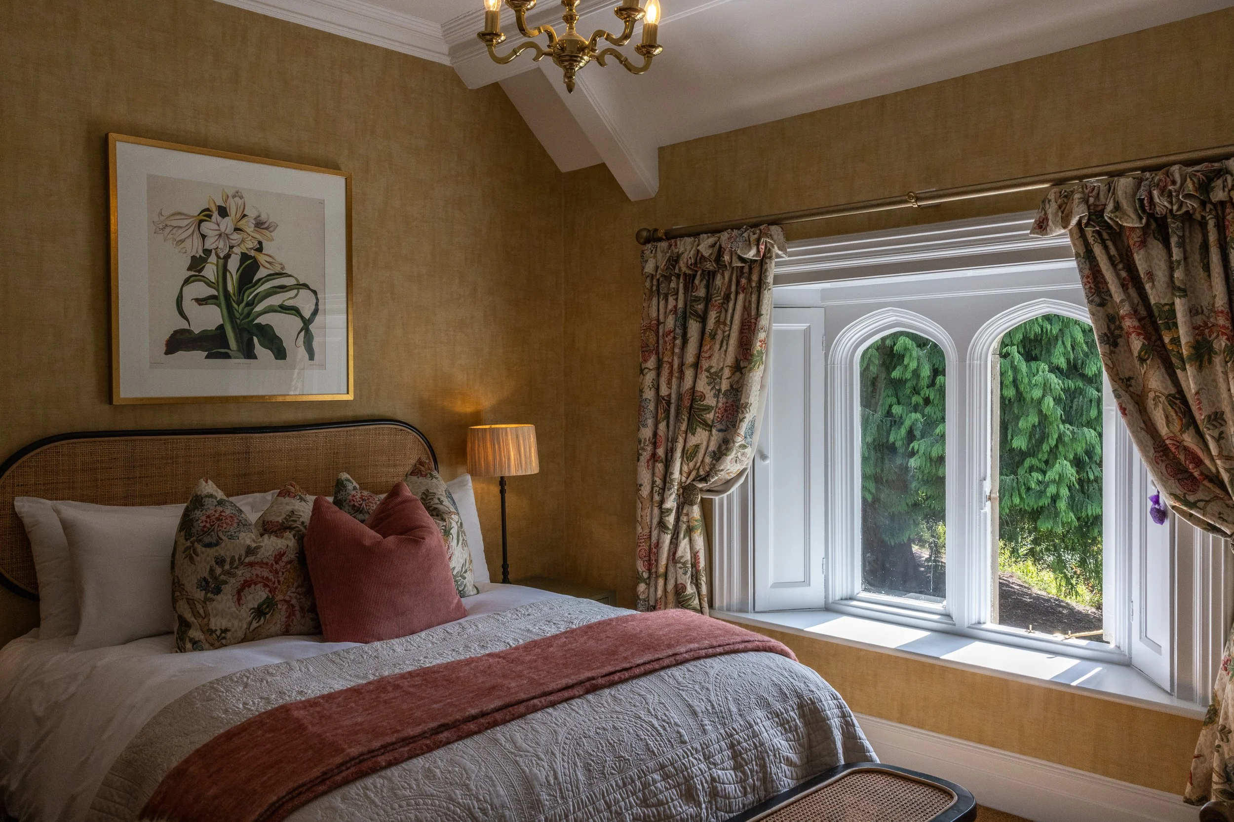 A bedroom with a large window, floral curtains, a bed with pillows and a quilt, a nightstand with a table lamp, a framed flower artwork on the wall, and a leafy view outside the window.