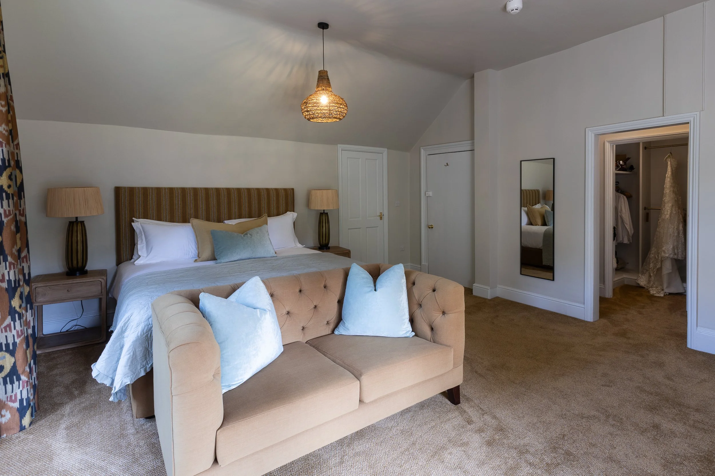 A bedroom with a beige tufted sofa at the foot of a bed with a striped beige headboard, beige and light blue pillows, and two matching bedside lamps. A mirror is on the wall next to the door to a walk-in closet with a wedding dress hanging inside.