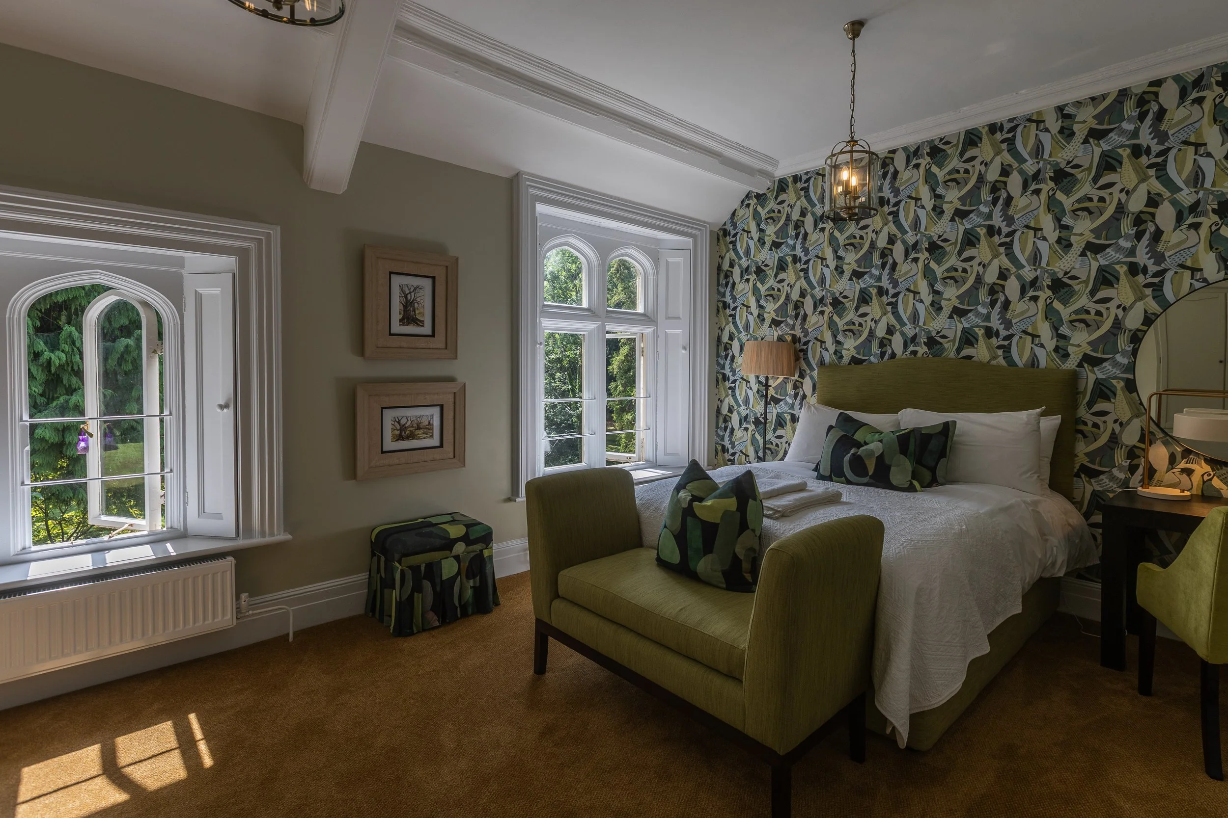 A bedroom with two large windows, a bed with green headboard and matching pillows, a green upholstered bench at the foot of the bed, a black nightstand with a lamp, patterned wallpaper behind the bed, and framed artwork on the beige wall. The room features a brown carpet and hanging chandelier.
