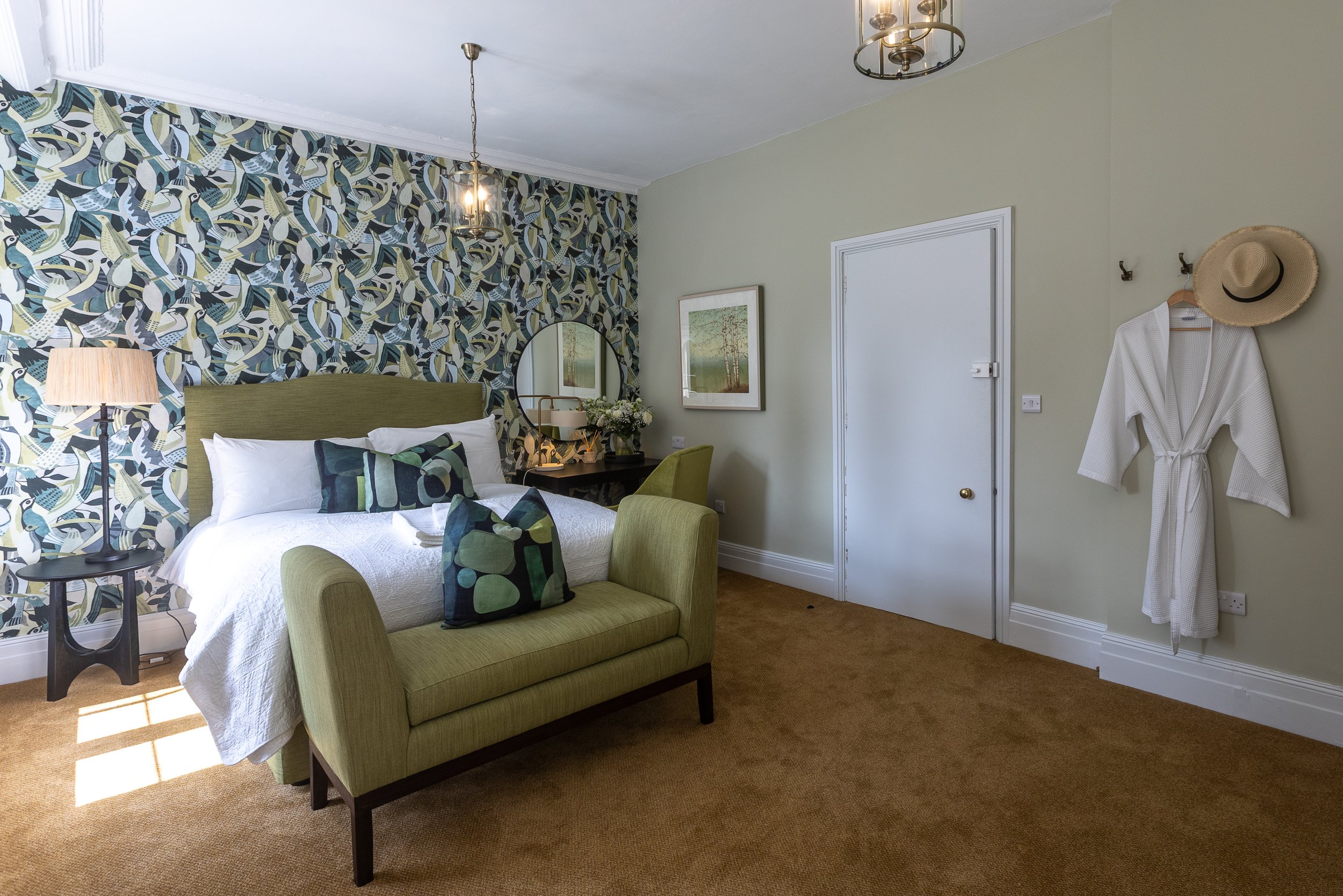 A cozy hotel room with a bed, green headboard, and decorative pillows. There is a patterned wallpaper behind the bed, a nightstand with a lamp, and a vanity or desk with a mirror. A white robe and straw hat hang on the wall, and framed artwork is on 