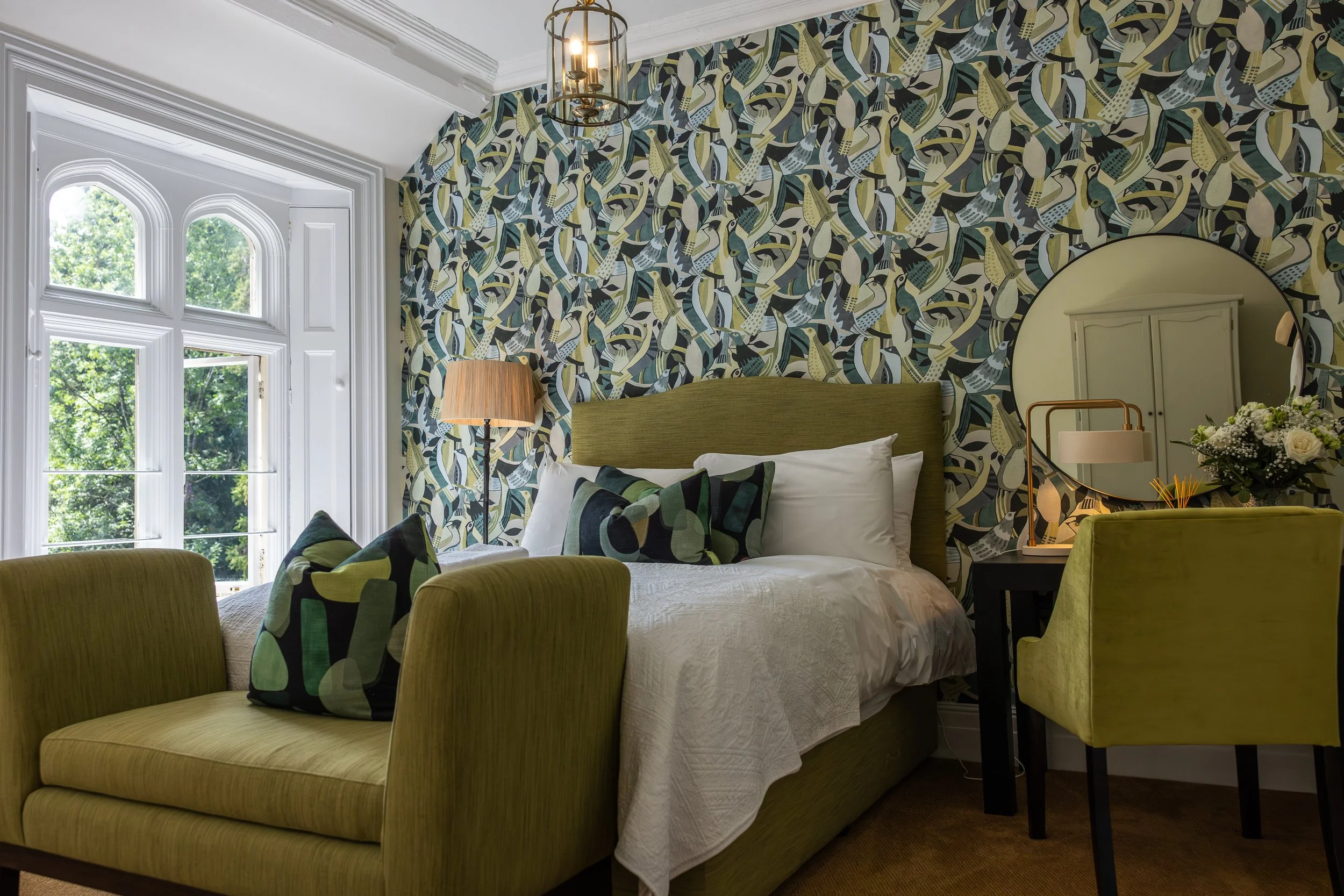 A bedroom with a green upholstered bed, white bedding, patterned pillows, and a matching green upholstered bench at the foot of the bed. The wall behind the bed features a colorful bird wallpaper. There is a window with rounded arches on the left, al