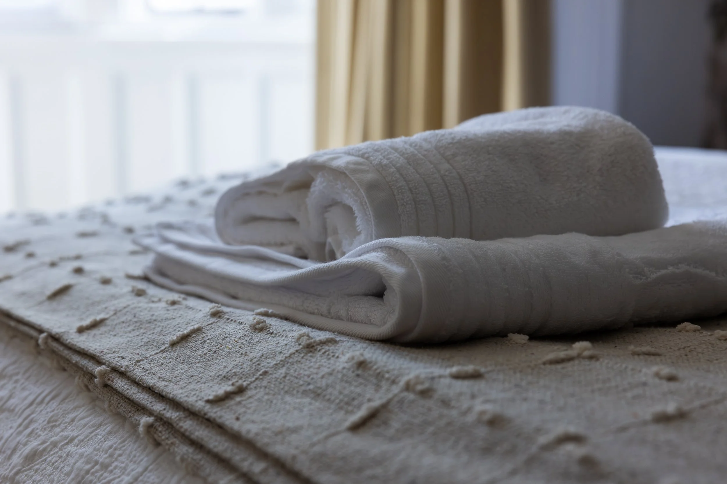 Two rolled white towels resting on a folded white towel on a bed, with light coming through the window in the background.