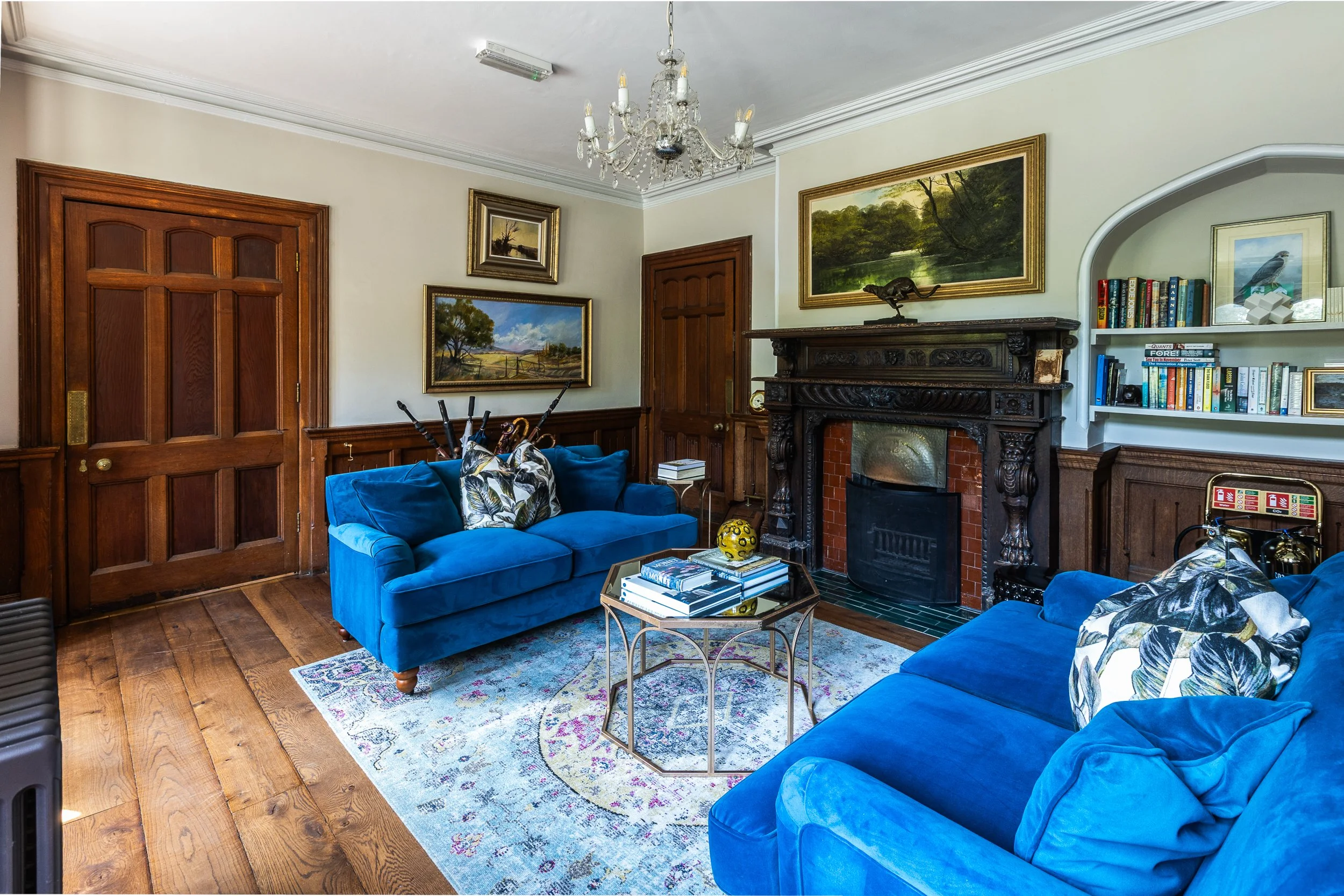 Living room with blue upholstered sofas, a decorative rug, a vintage fireplace, wooden wall paneling, framed landscape paintings, a chandelier, and a bookshelf filled with books.