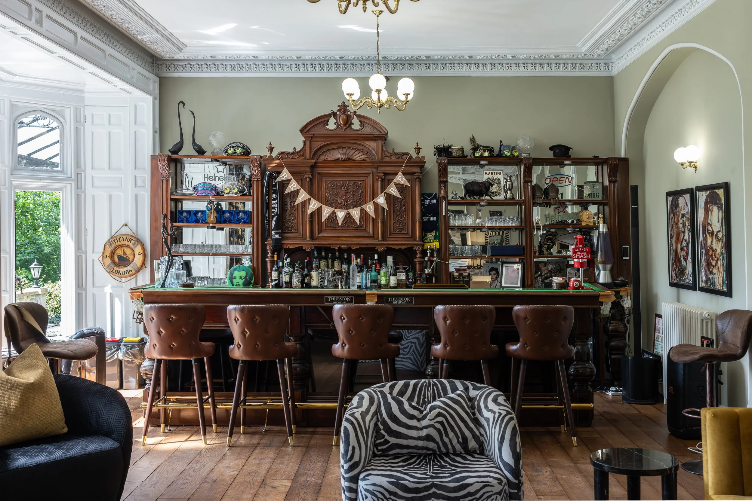 An ornate home bar with a wooden top and a carved wooden back panel. There are five brown bar stools in front of the bar. Shelves behind the bar are filled with bottles, glasses, and various decorations such as a black pig, a sculpture, and signs. A banner spelling 'BRIDE TO BE' hangs above the bar. To the left, there is a window with white trim, a black armchair with a beige pillow, and a chair with a zebra print cover in front. On the right side, there are two framed pieces of artwork, a black speaker, and a small round black table. The room has a wooden floor, white crown molding, and a chandelier hanging from the ceiling.