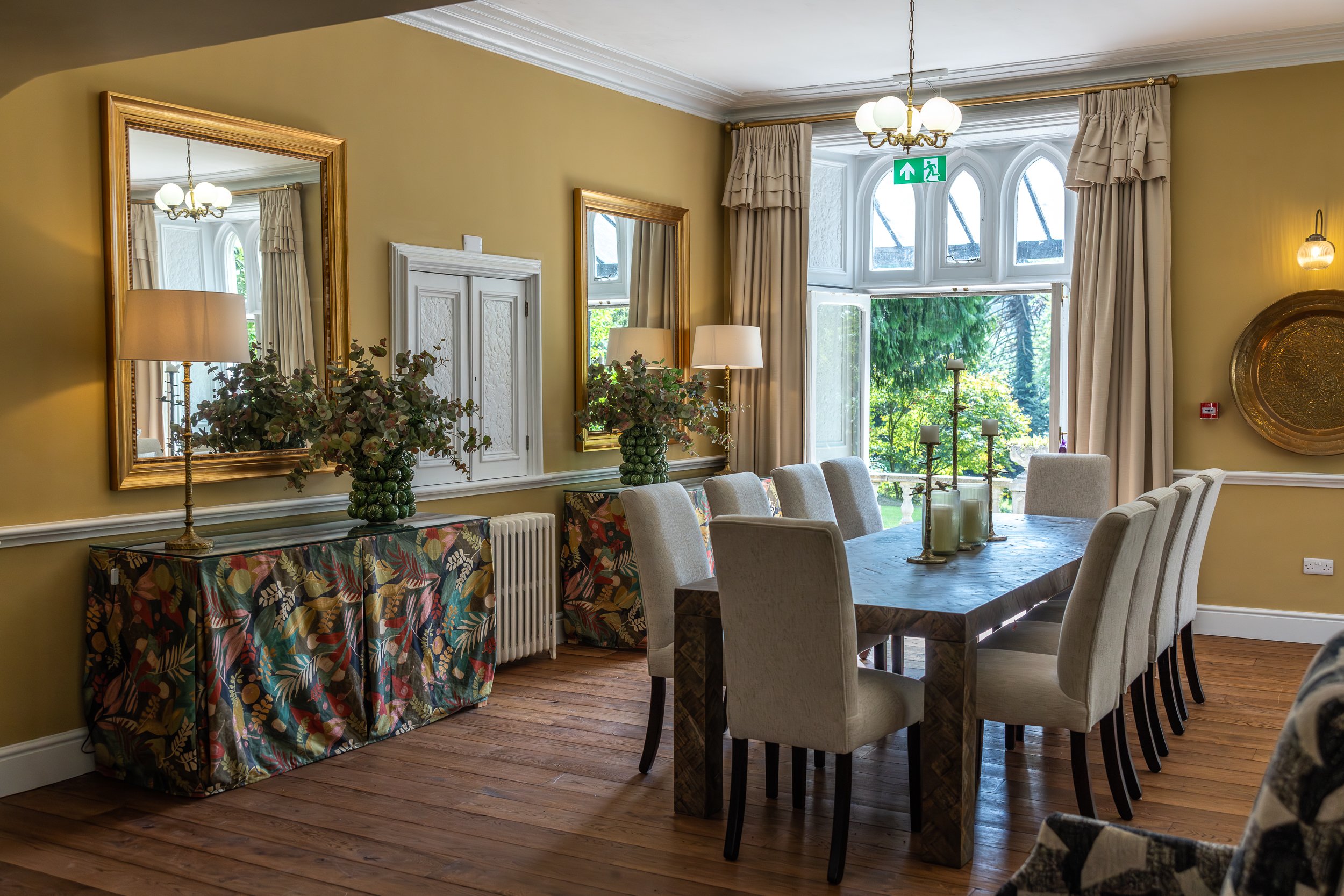 A dining room with a wooden table surrounded by beige upholstered chairs, a large window with beige curtains, two large mirrors with gold frames, and floral decorations on a console table.