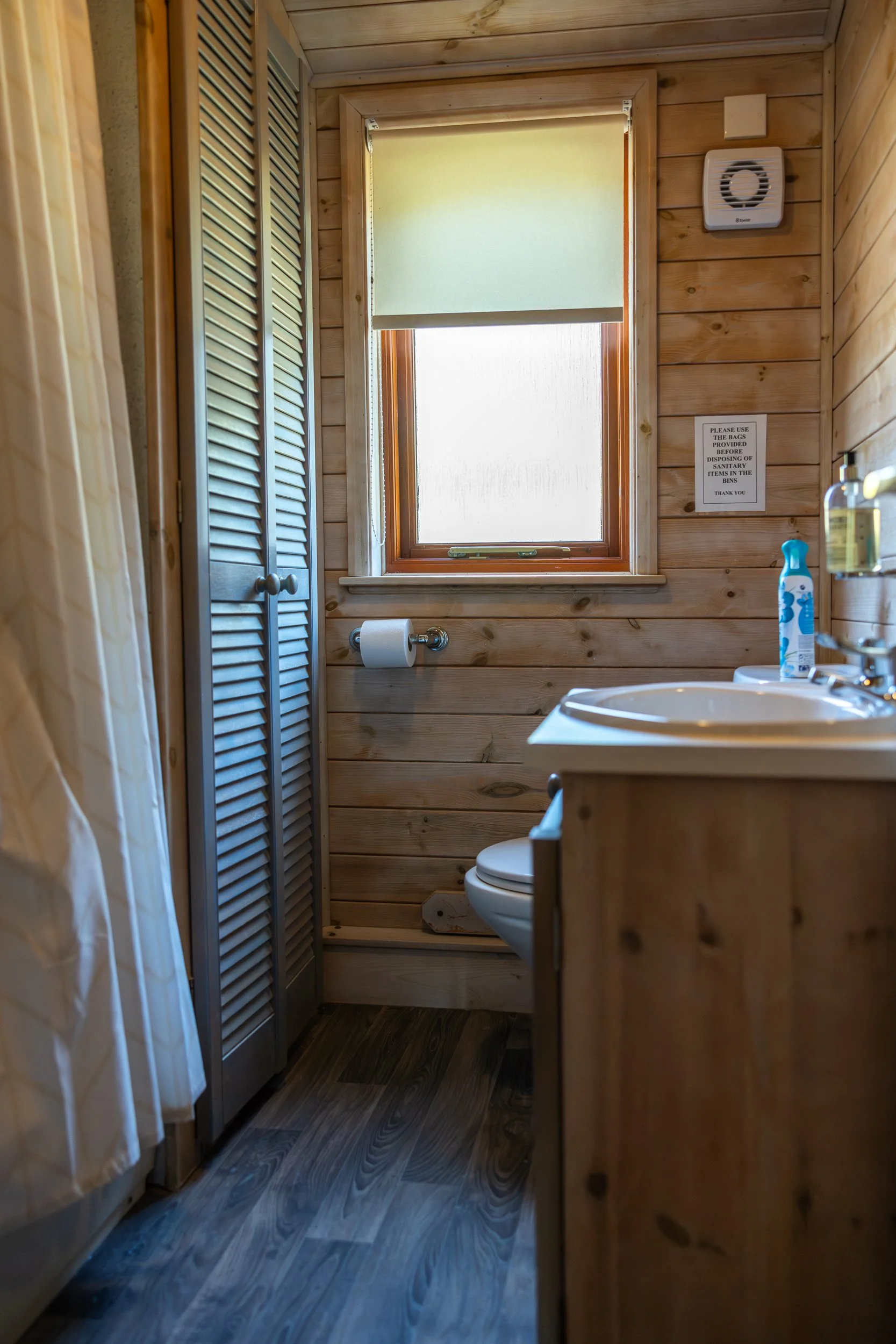 Small bathroom with wooden walls, a small window with a roller shade, a toilet, a sink, a soap dispenser, and a sign on the wall.